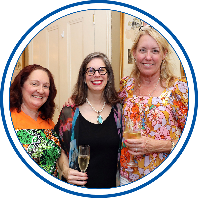 Three women smiling at a social gathering, one holding a glass of champagne, in an indoor setting with a beige door and wall decorations in the background.