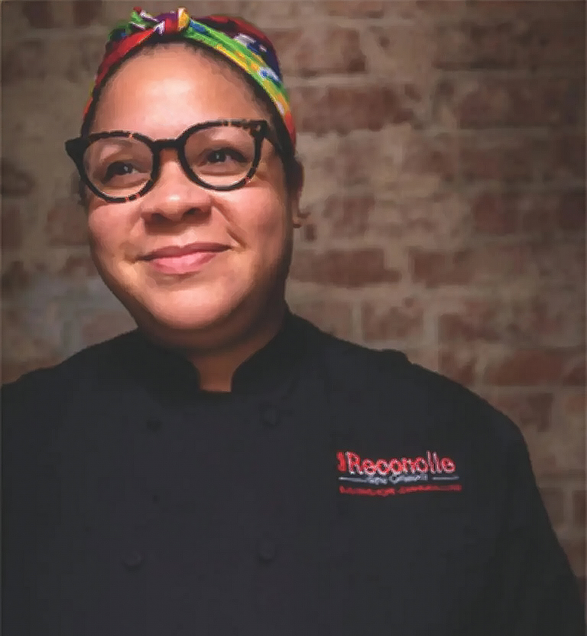 A woman wearing black chef's coat with red embroidery, black glasses, and a colorful headband, standing against a brick wall.