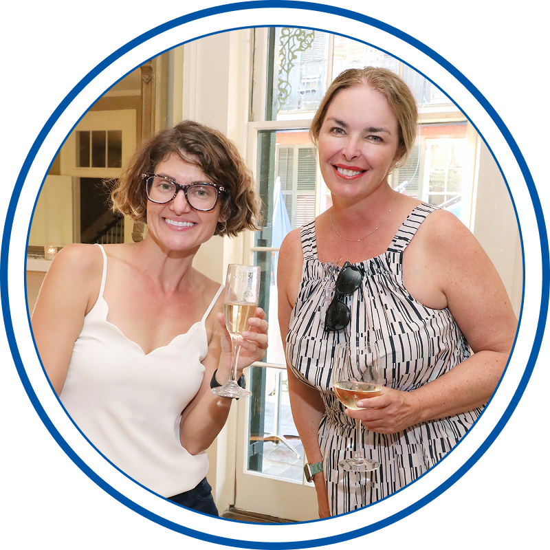Two women standing indoors, smiling, holding glasses of white wine, at a social gathering.