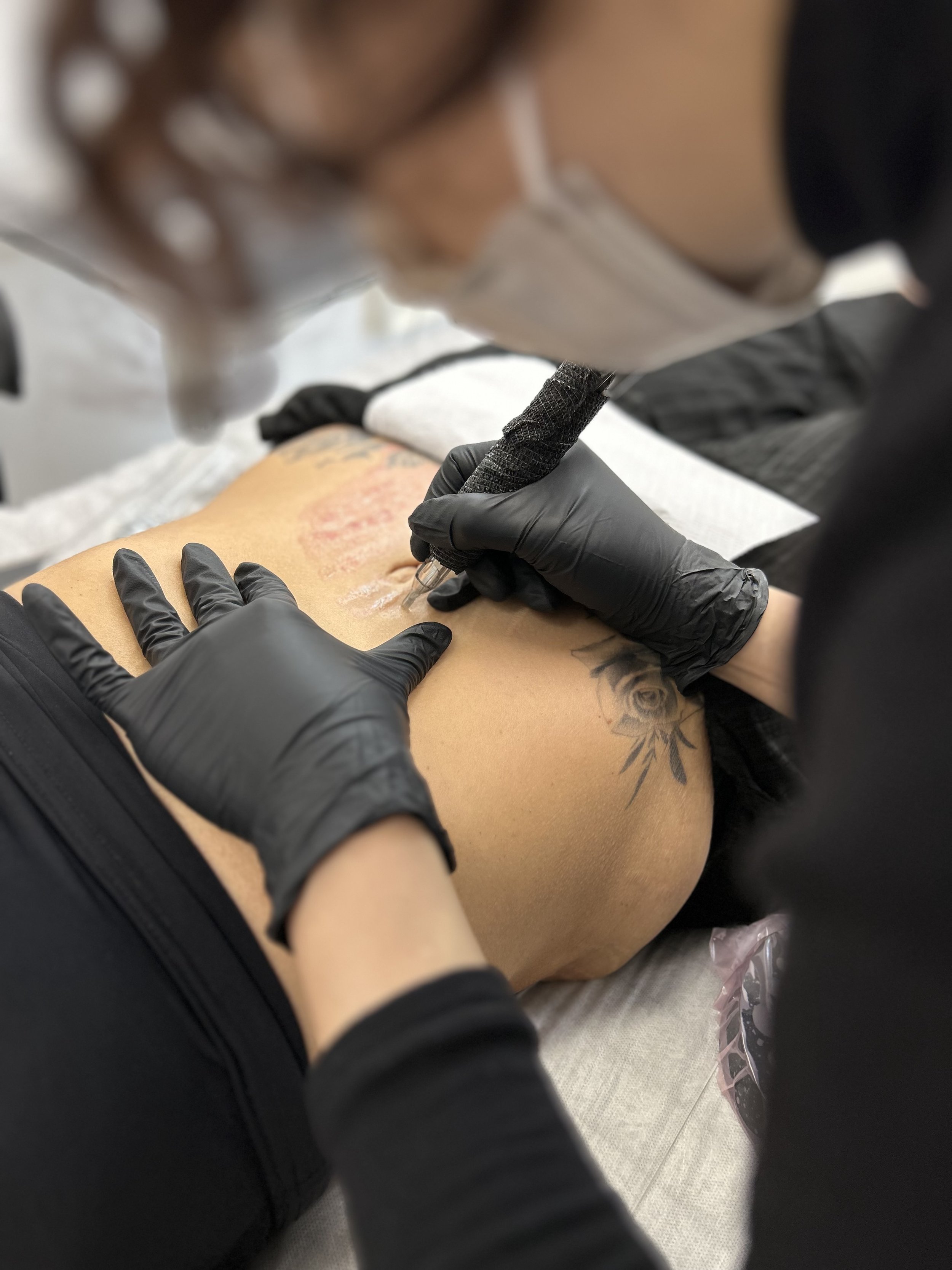 A person receiving a tattoo on their side or ribcage area from a tattoo artist. The artist is wearing black gloves and is using a tattoo machine. The person's skin appears red and irritated from the tattooing process, and there is a floral tattoo near the new tattoo.
