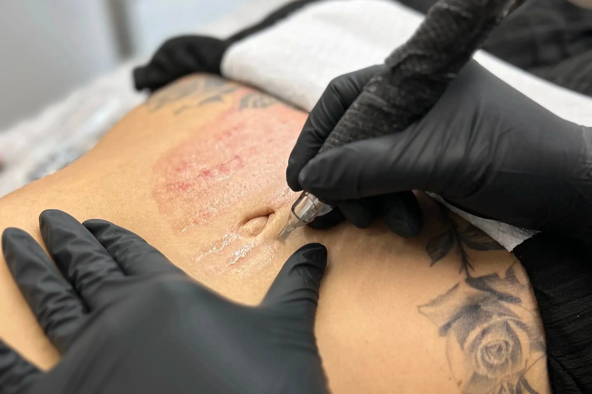 A person with tattoos receiving a tattoo on their arm from a tattoo artist wearing black gloves.
