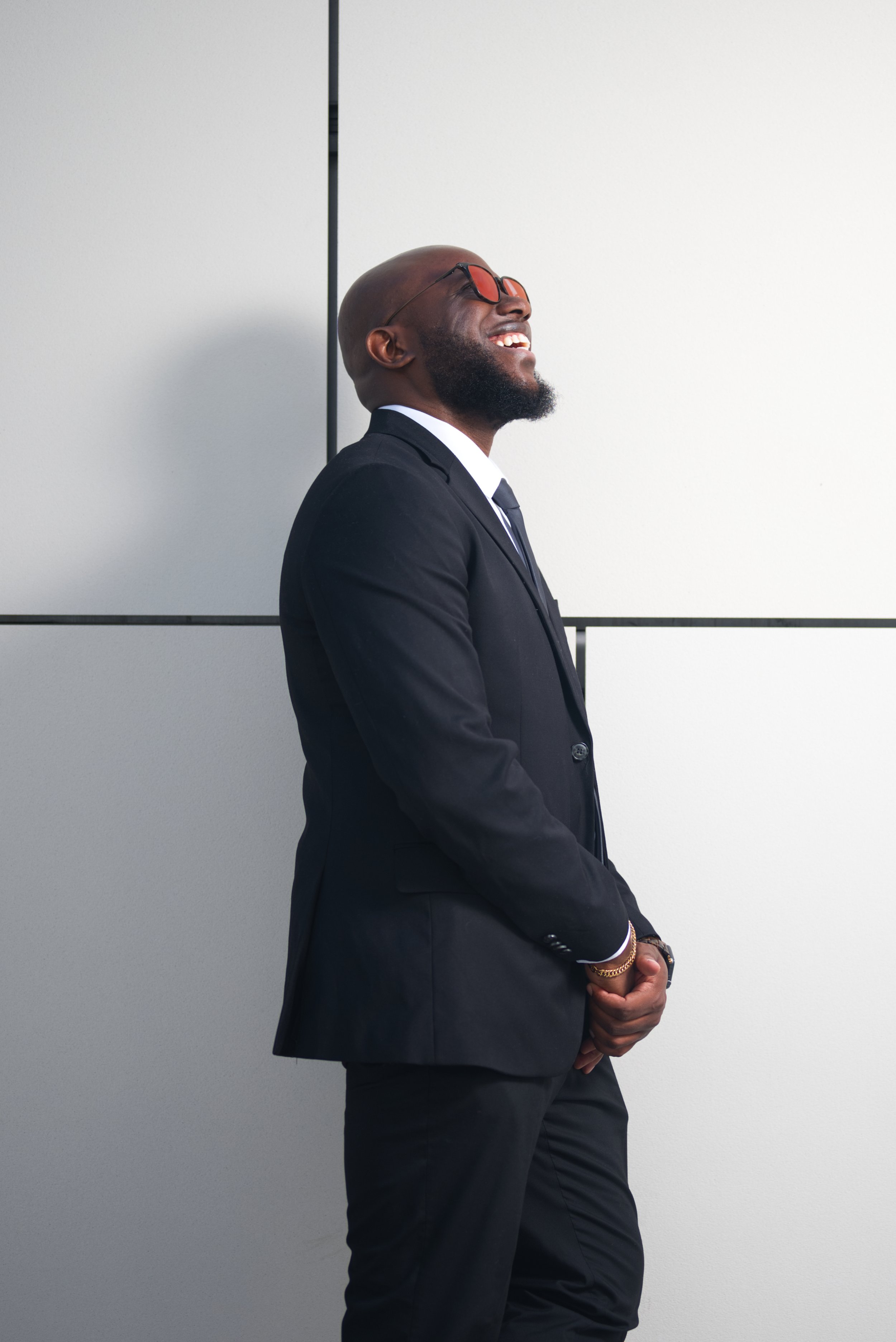 A man dressed in a black suit, white shirt, and black tie, smiling with glasses and a beard, standing side profile against a plain light gray background.