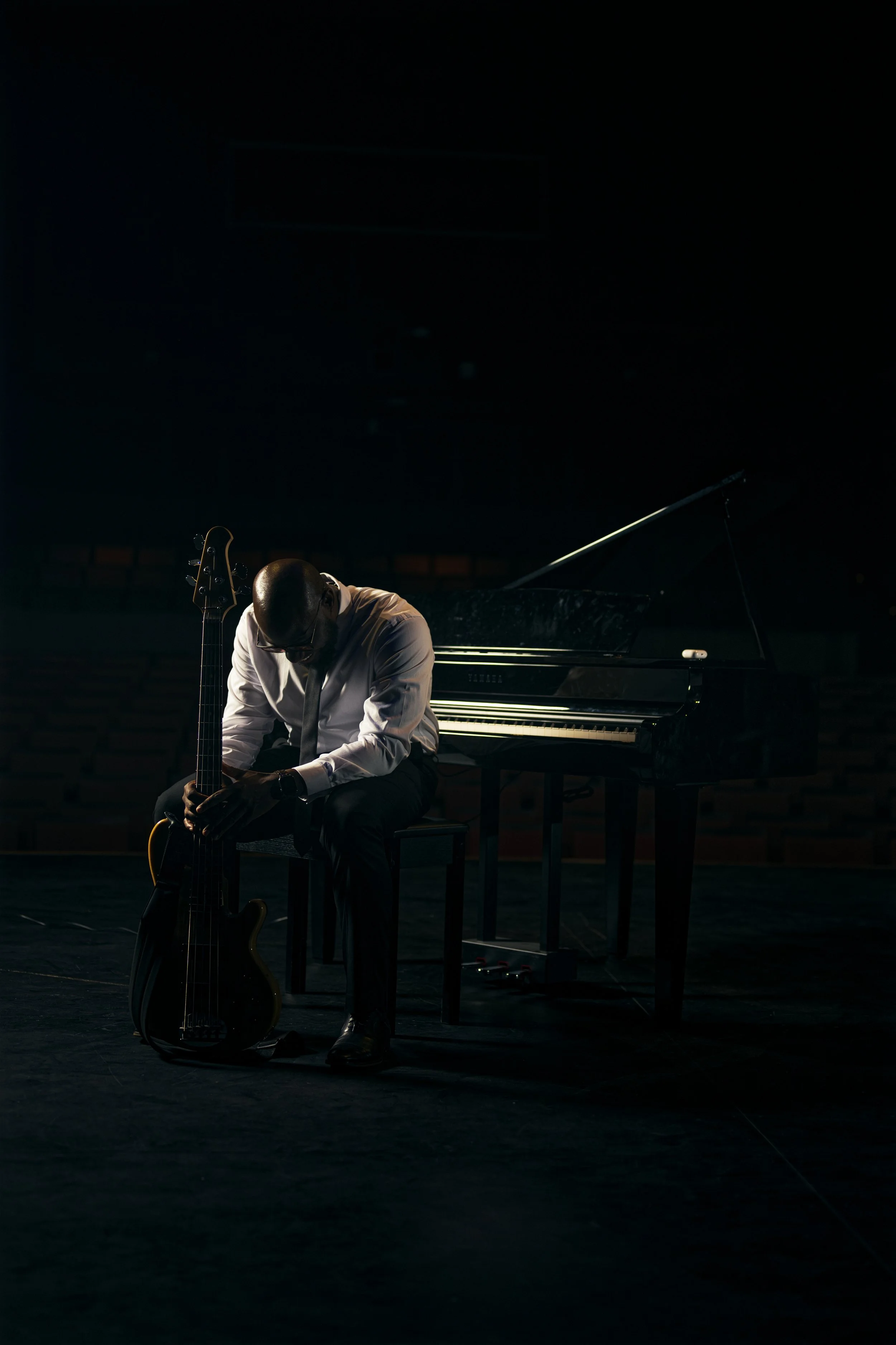 A man in a white shirt and tie sits on a chair in a dark room, holding an electric guitar. A grand piano is behind him, and a music stand is to his right. The setting appears to be a stage or rehearsal space.