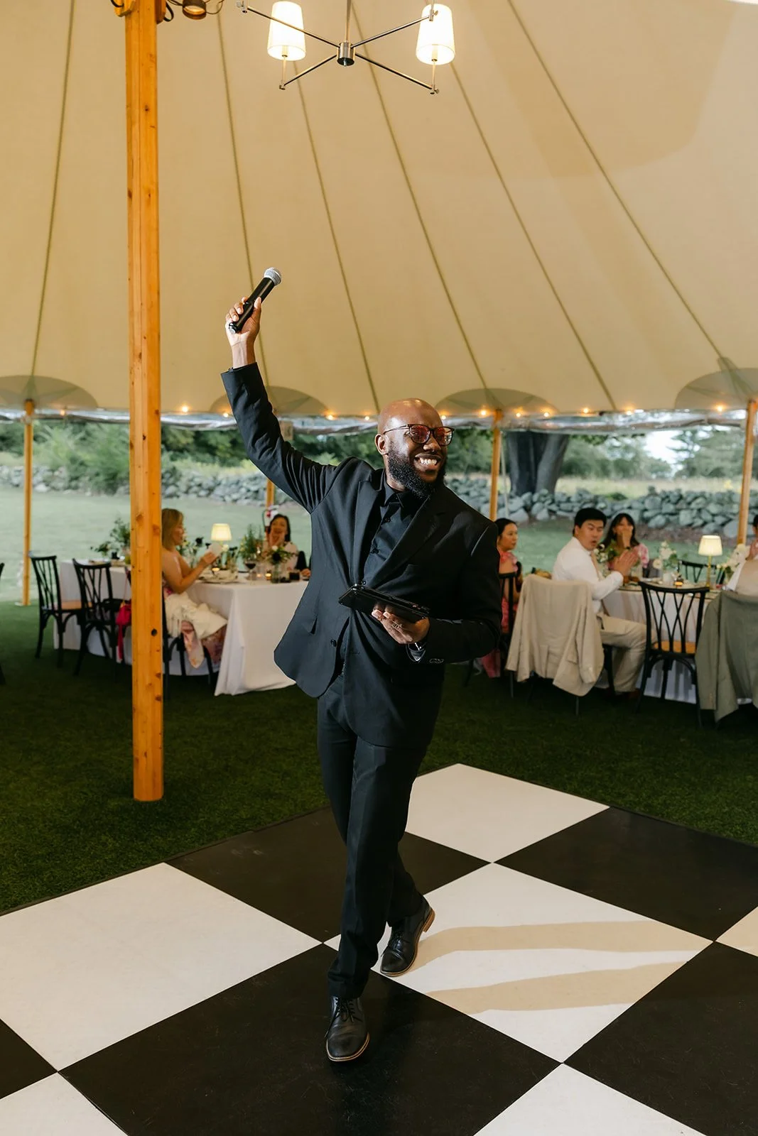 Man in a black suit and glasses, holding a microphone and a tablet, dancing on a black and white checkered dance floor inside a large tent at a formal event with seated guests in the background.