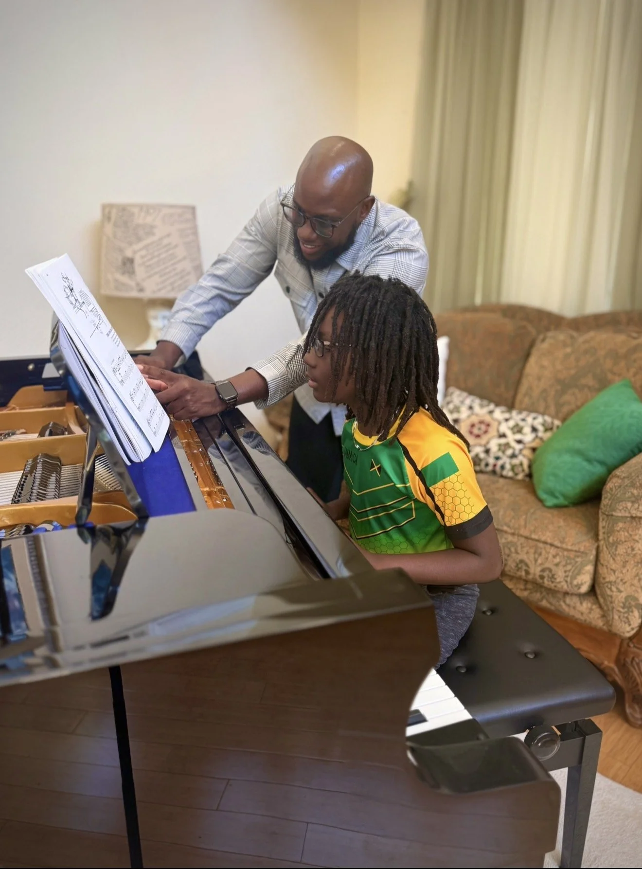 A man and a young boy are sharing a moment at a piano, with the man guiding the boy's hands over the keys, in a cozy living room setting.