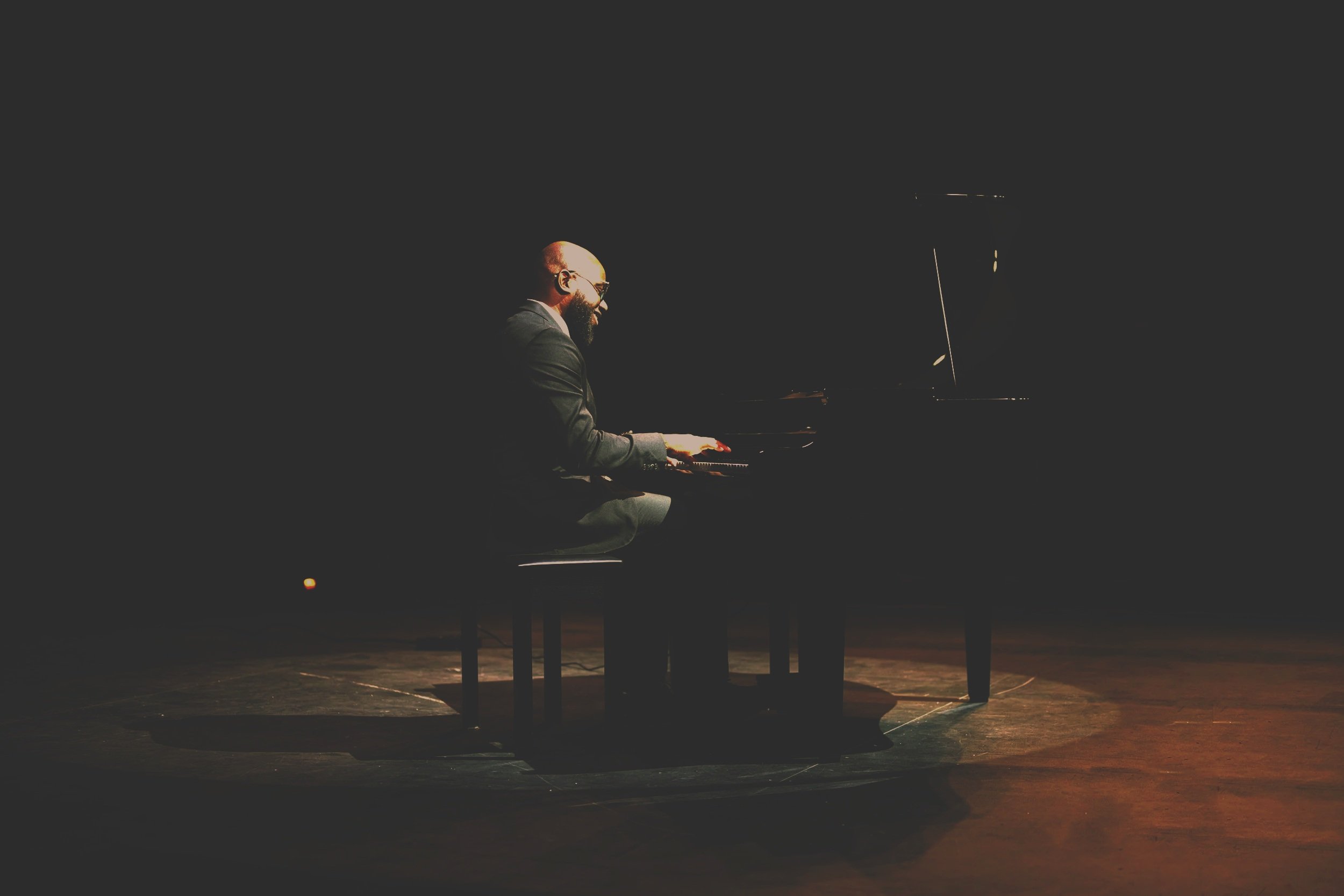 A man with a beard and glasses playing a grand piano on a dark stage.