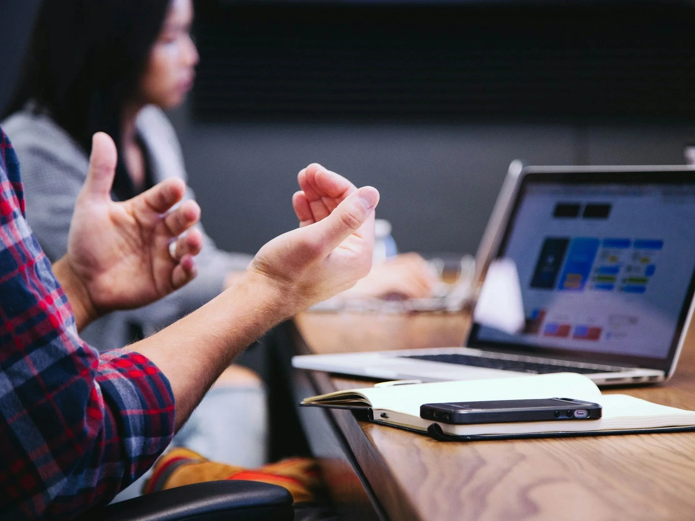 A person in a red and blue plaid shirt is gesturing with their hand in a discussion or presentation during a meeting. There is a laptop and a notebook on the table in front of them.