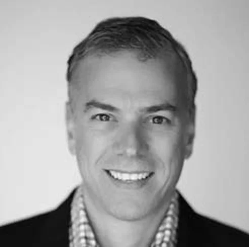 Black-and-white headshot of a smiling man wearing a checkered shirt and blazer.