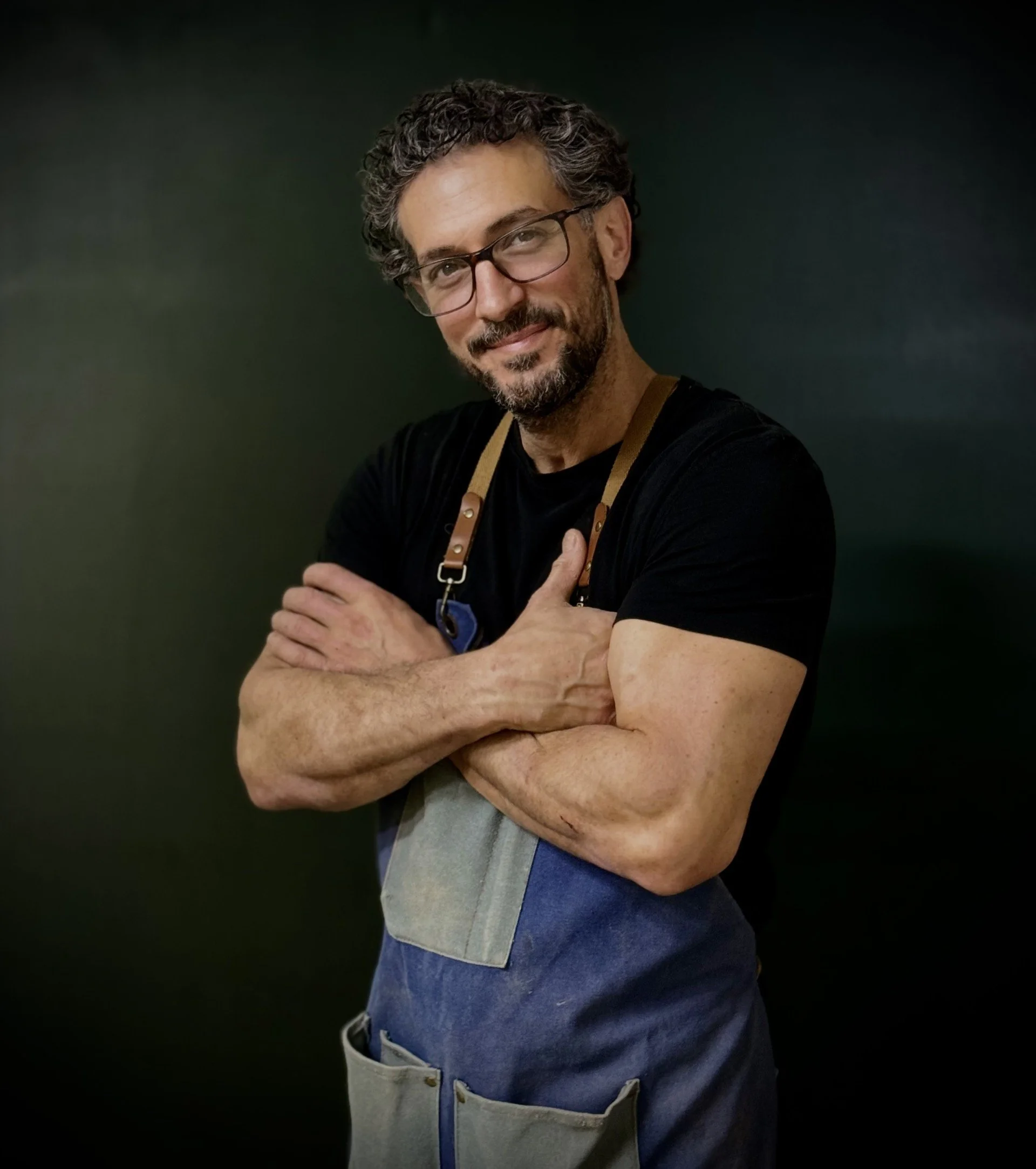 Handsome man with curly gray hair, beard, and glasses, wearing a black shirt and an apron, standing against a dark background.