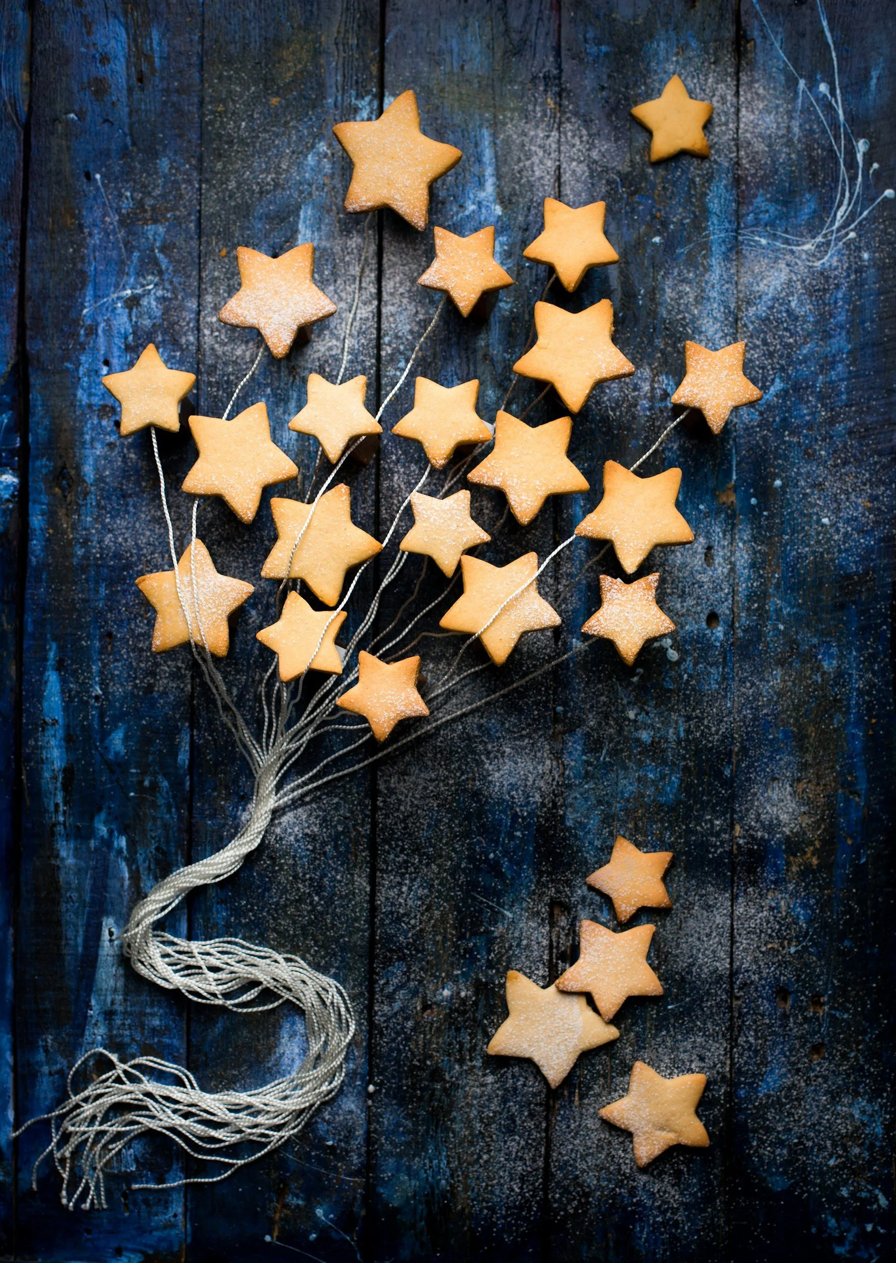 Star-shaped cookies dusted with powdered sugar arranged on a dark blue wooden surface, with white string decorations and scattered powdered sugar for a festive holiday appearance.