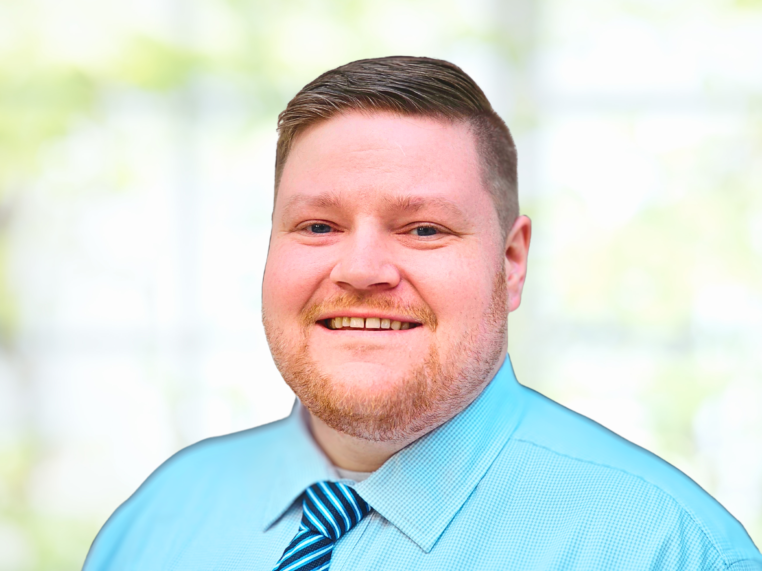 Close-up of a man with short brown hair, blue eyes, and a light beard, smiling, wearing a light blue shirt and a striped blue tie, with a blurred outdoor background.