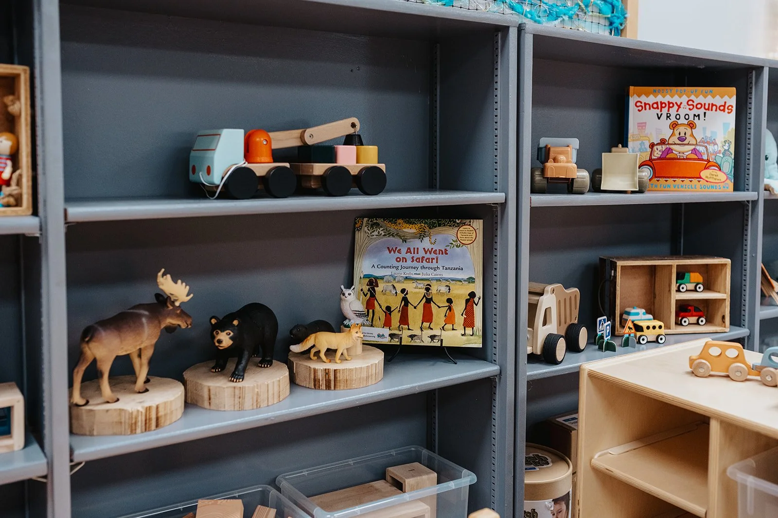 Wooden children's toys on grey shelves, including animal figurines, toy vehicles, and picture books.