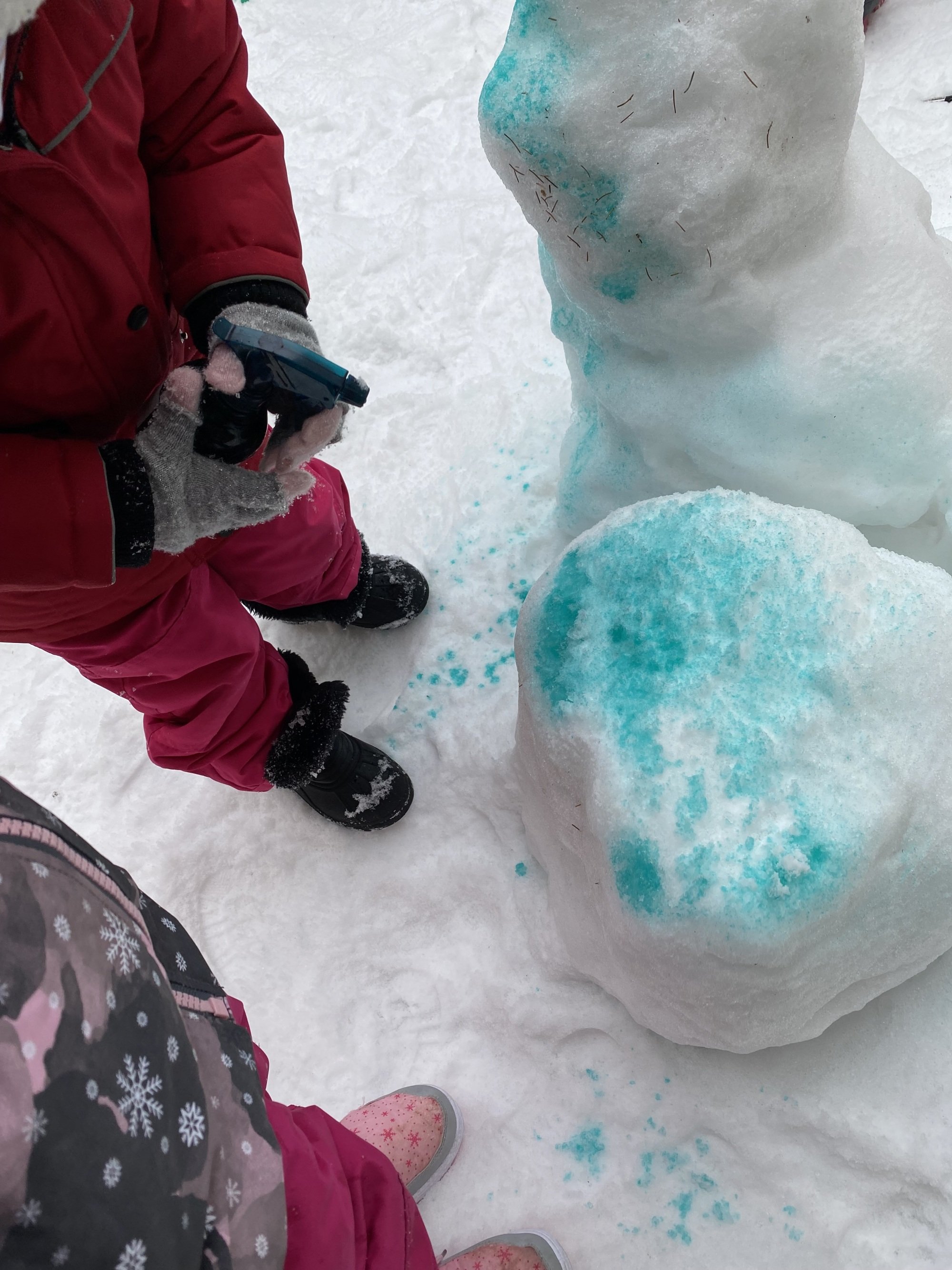 Children dressed in winter clothing playing in the snow and building a snowman, with blue paint or dye on the snow.