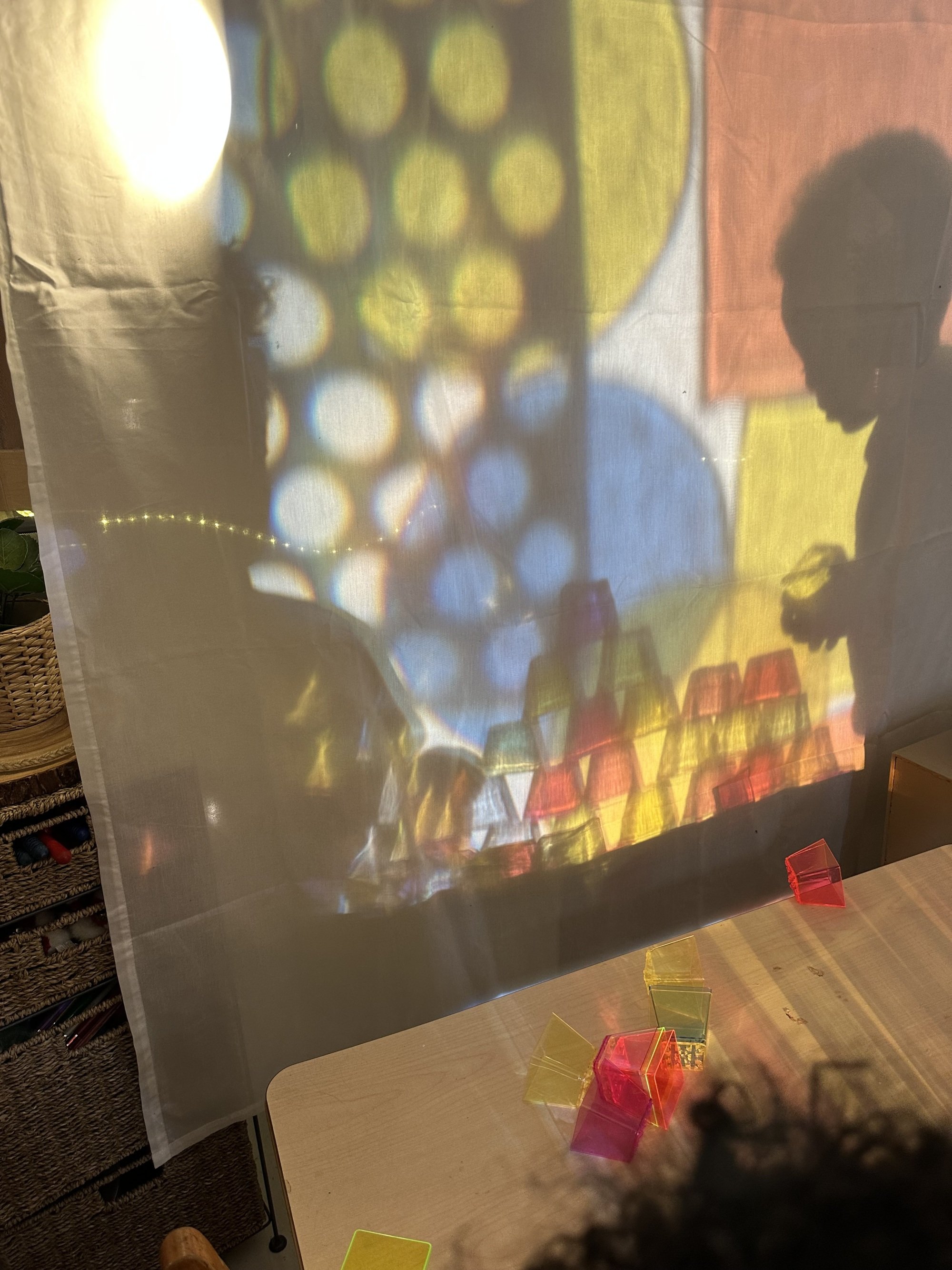 Colorful geometric shapes and light patterns projected onto a semi-transparent surface, with several small colorful translucent cubes on a light wooden table in the foreground.