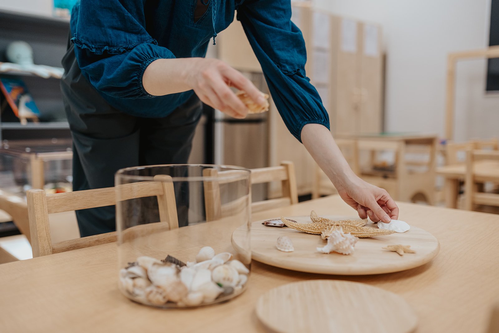 Person arranging seashells and starfish on a wooden table, with a glass bowl of seashells in the foreground.
