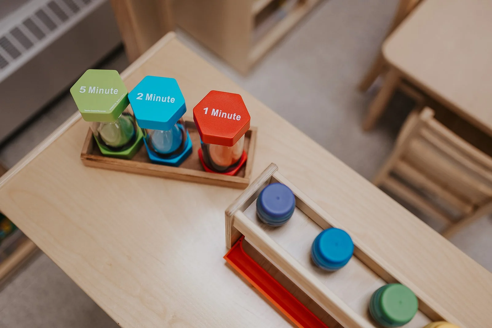 Colorful sand timers labeled 5 minutes, 2 minutes, and 1 minute on a wooden table in a classroom.