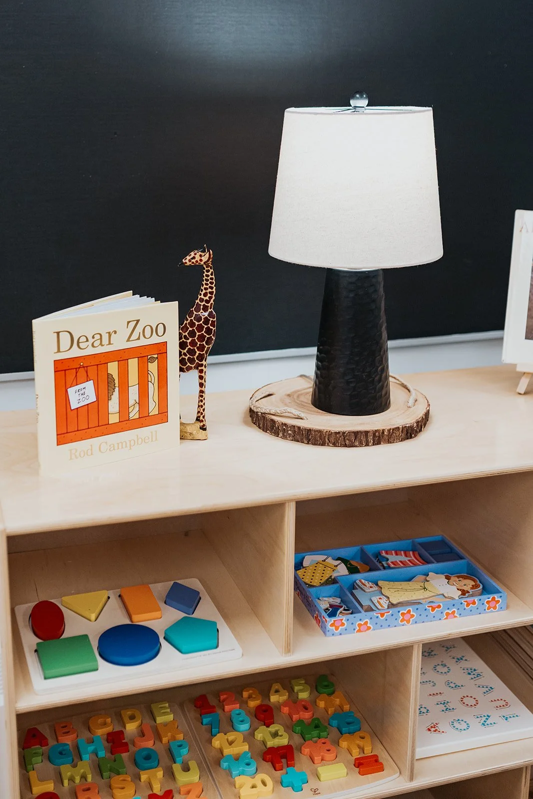 A wooden table with children’s educational toys and a children’s book titled 'Dear Zoo' by Rod Campbell. The table has a black lampshade on a textured black lamp base, a giraffe figurine, and various color and number puzzles.