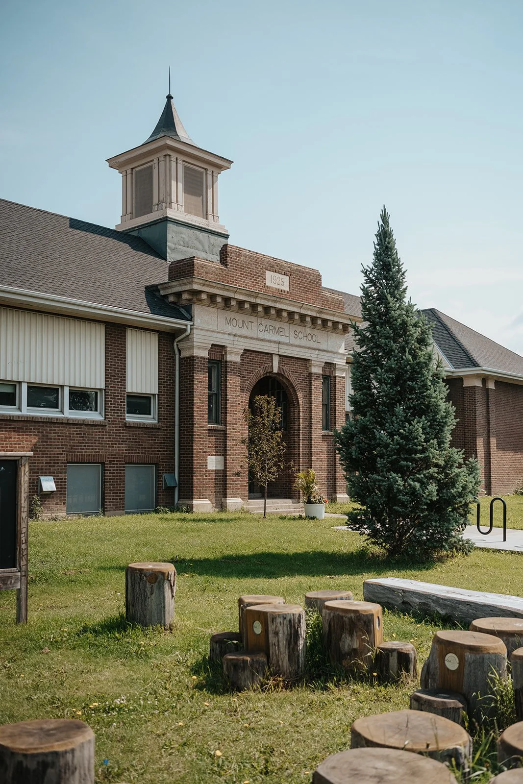 A brick school building with a tower, sign reading 'Mount Carmel School' and trees in front, in a grassy area with wooden stumps.