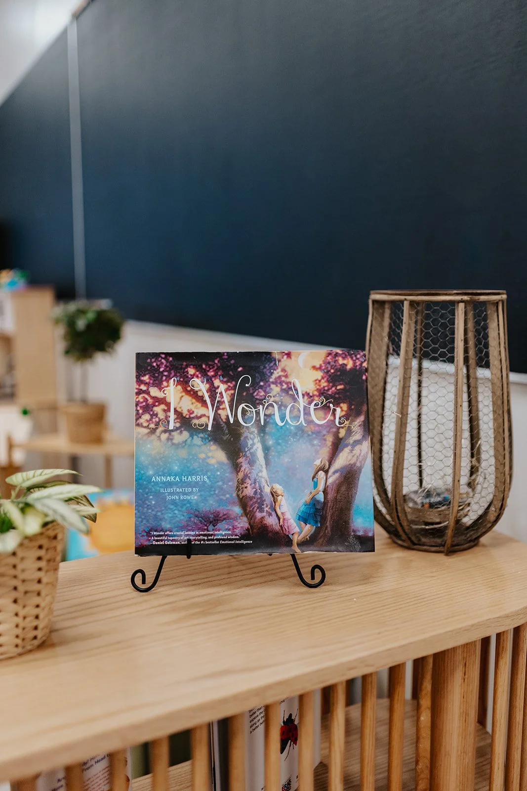 A children's book titled 'I Wonder' by Annaka Harris, illustrated by John Rowe, is displayed on a small black stand on a wooden table. The cover features an illustration of two children reaching out to a glowing, sparkling tree in a colorful, fantastical forest scene. To the right of the book, there is a decorative tall wire basket with a wooden frame. In the background, there are shelves with various objects and a dark blue wall.