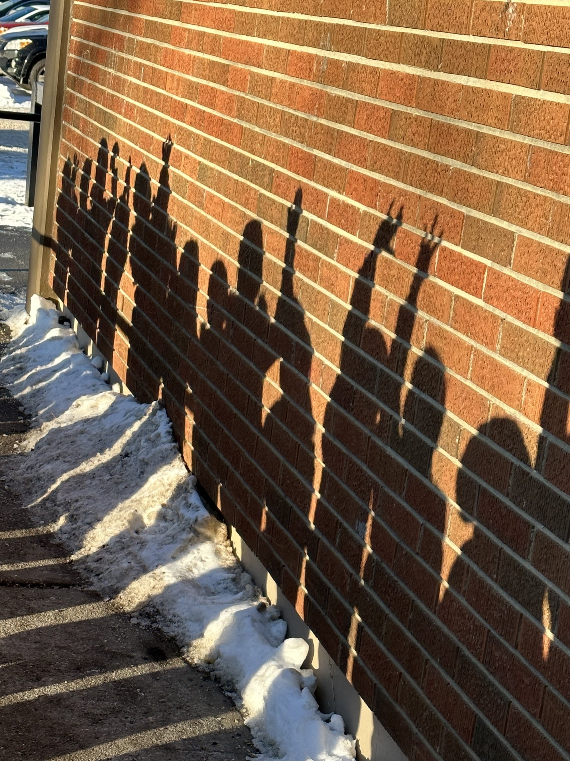 Shadow of several people cast on a brick wall with snow and a parking lot in the background.