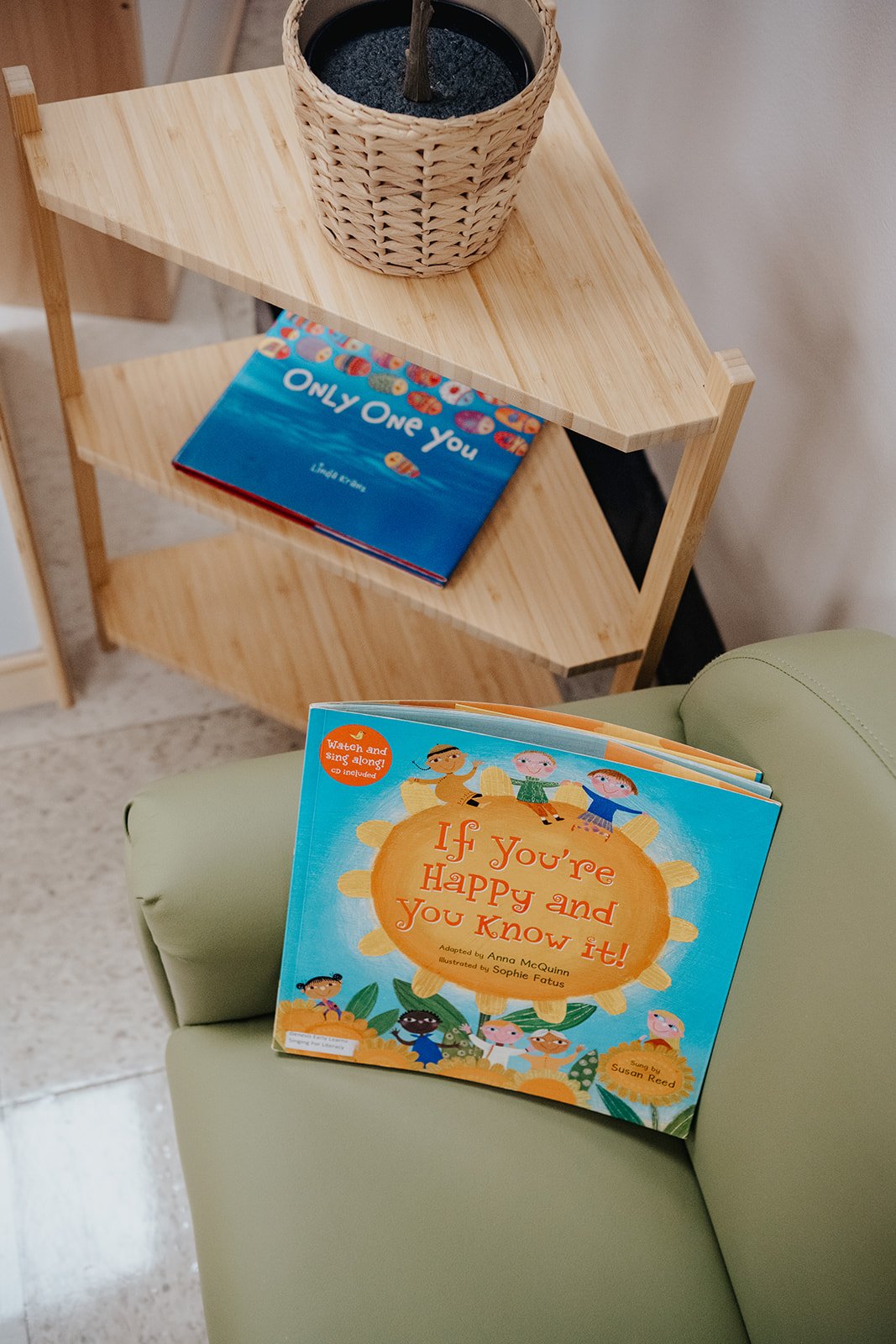 A small wooden corner shelf holding a woven basket with a plant or object inside and a children's book titled 'Only One You'. An open children's book titled 'If You're Happy and You Know It!' is resting on a green chair.