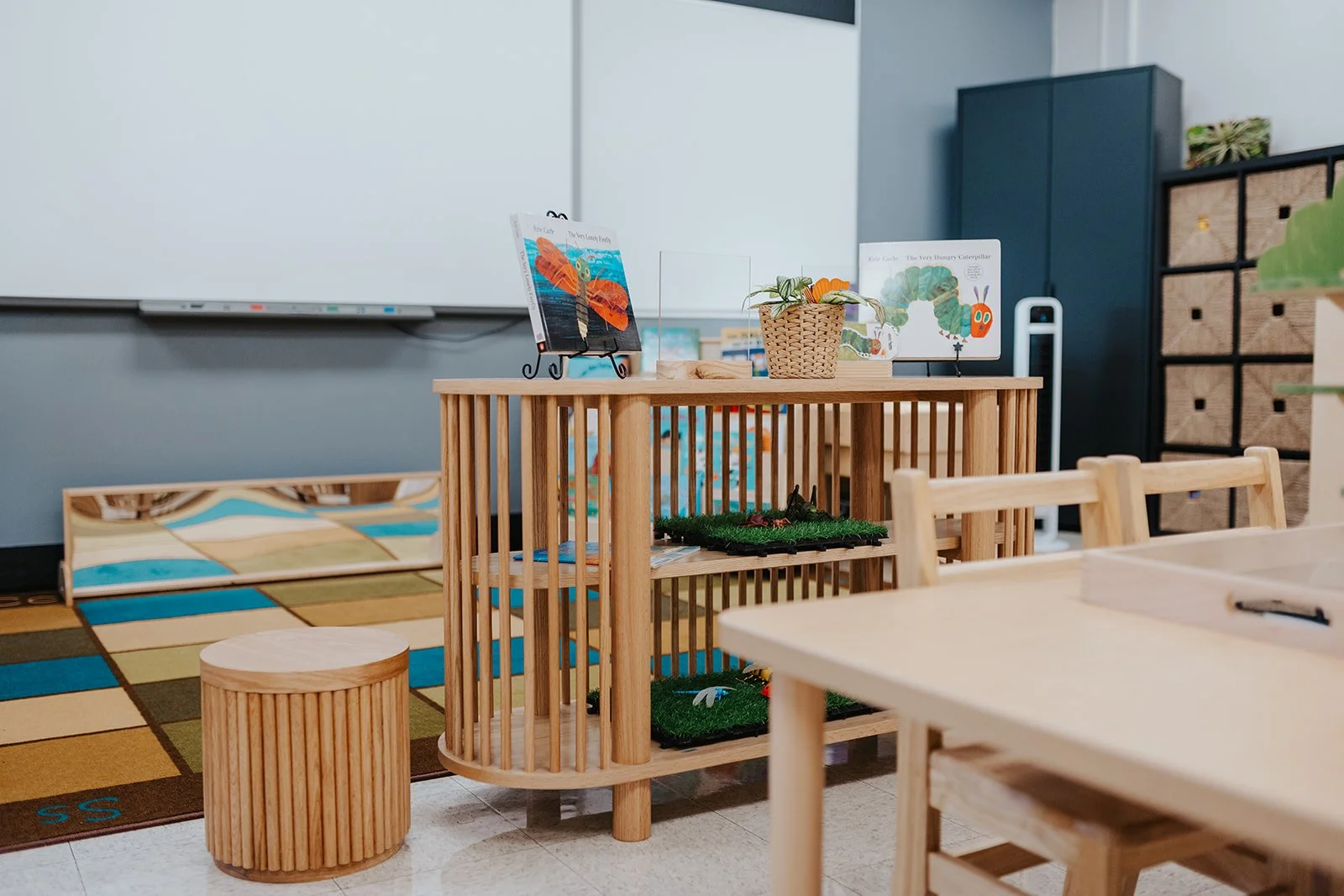 A classroom corner with a wooden shelving unit, children’s books, plush toys, and decor, with a whiteboard and storage cabinets in the background.