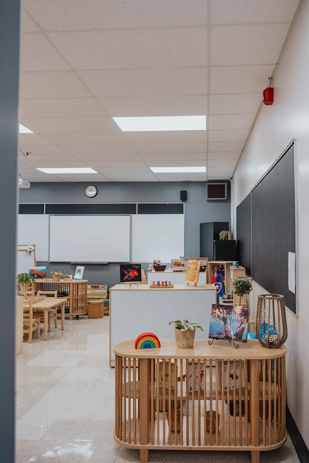 Interior of a classroom with small wooden tables and chairs, display boards, and educational decorations.