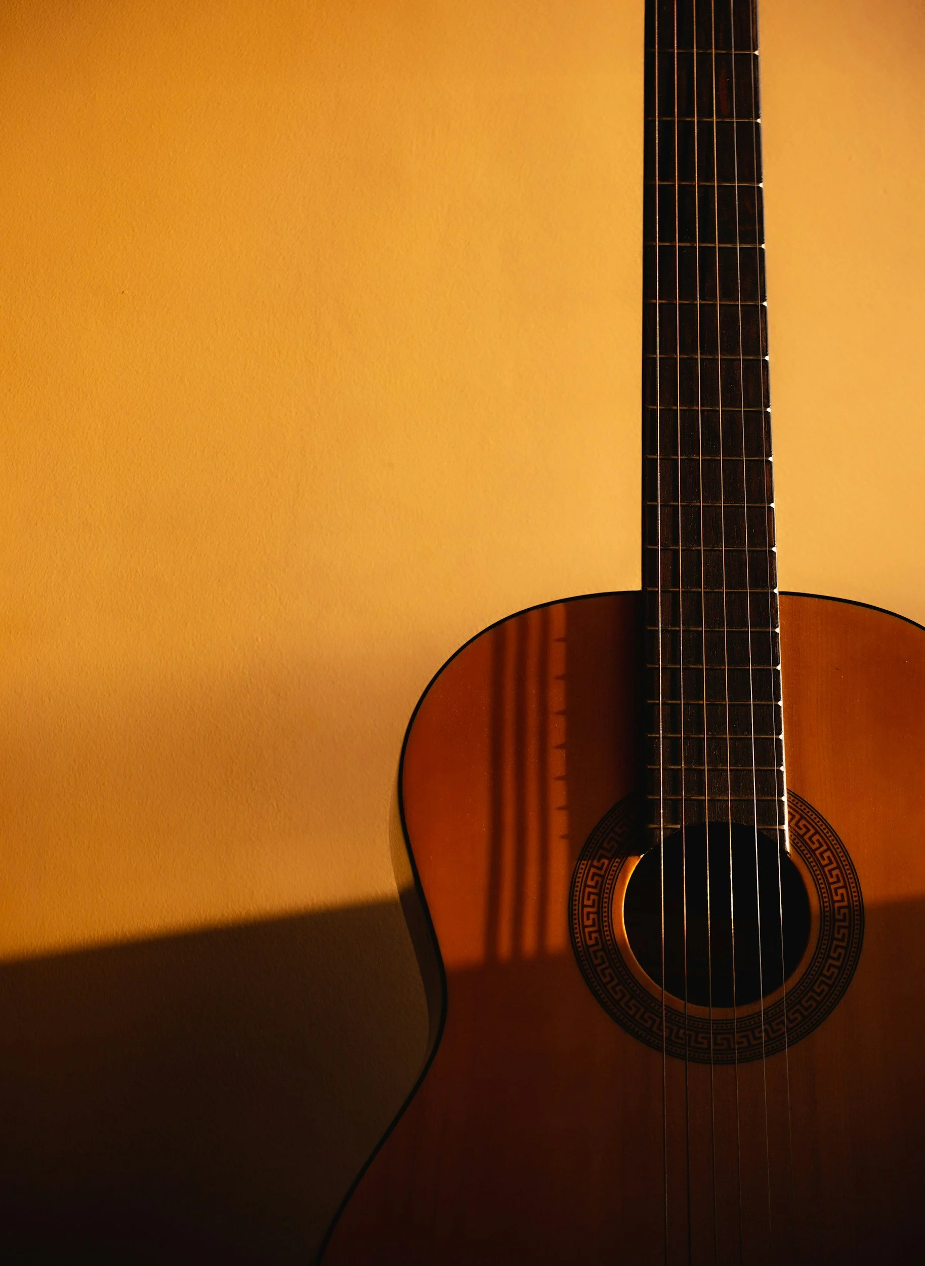 Close-up of a classical acoustic guitar against a yellow wall, showing part of the guitar body, neck, and strings.