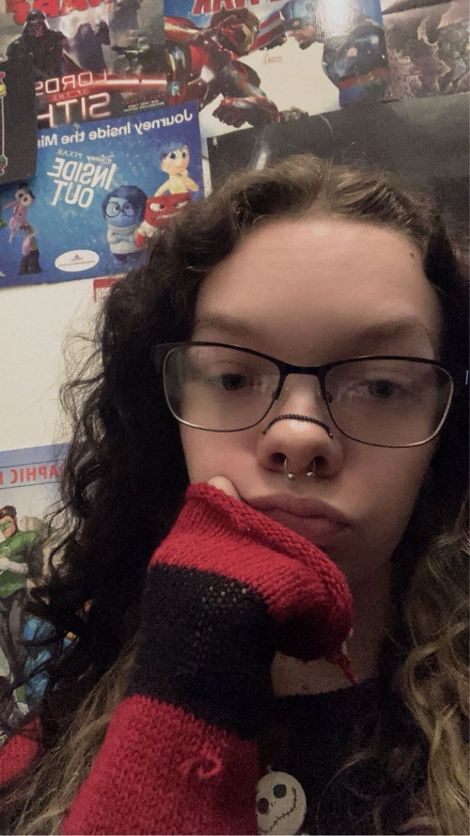 Close-up of a young person with long, curly hair, glasses, and a septum piercing, wearing a red and black striped glove with their hand resting on their face, in front of a wall decorated with posters from animated movies and superhero films.