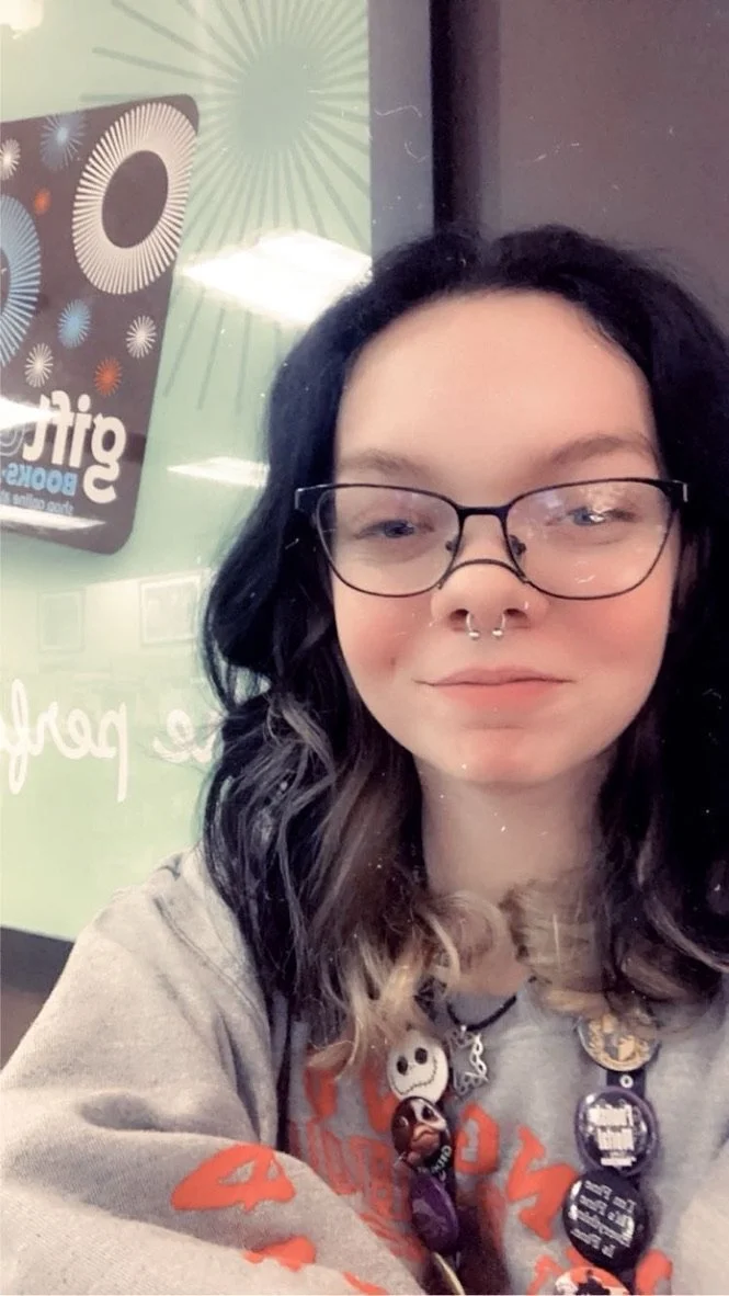 A young woman with wavy dark hair with blonde tips, glasses, and a septum piercing, smiling in a selfie. She is wearing a gray sweatshirt with orange letters and multiple buttons or pins on her chest area. Behind her, there is a glass window or door with a sign and some colorful, firework-like designs in the background.