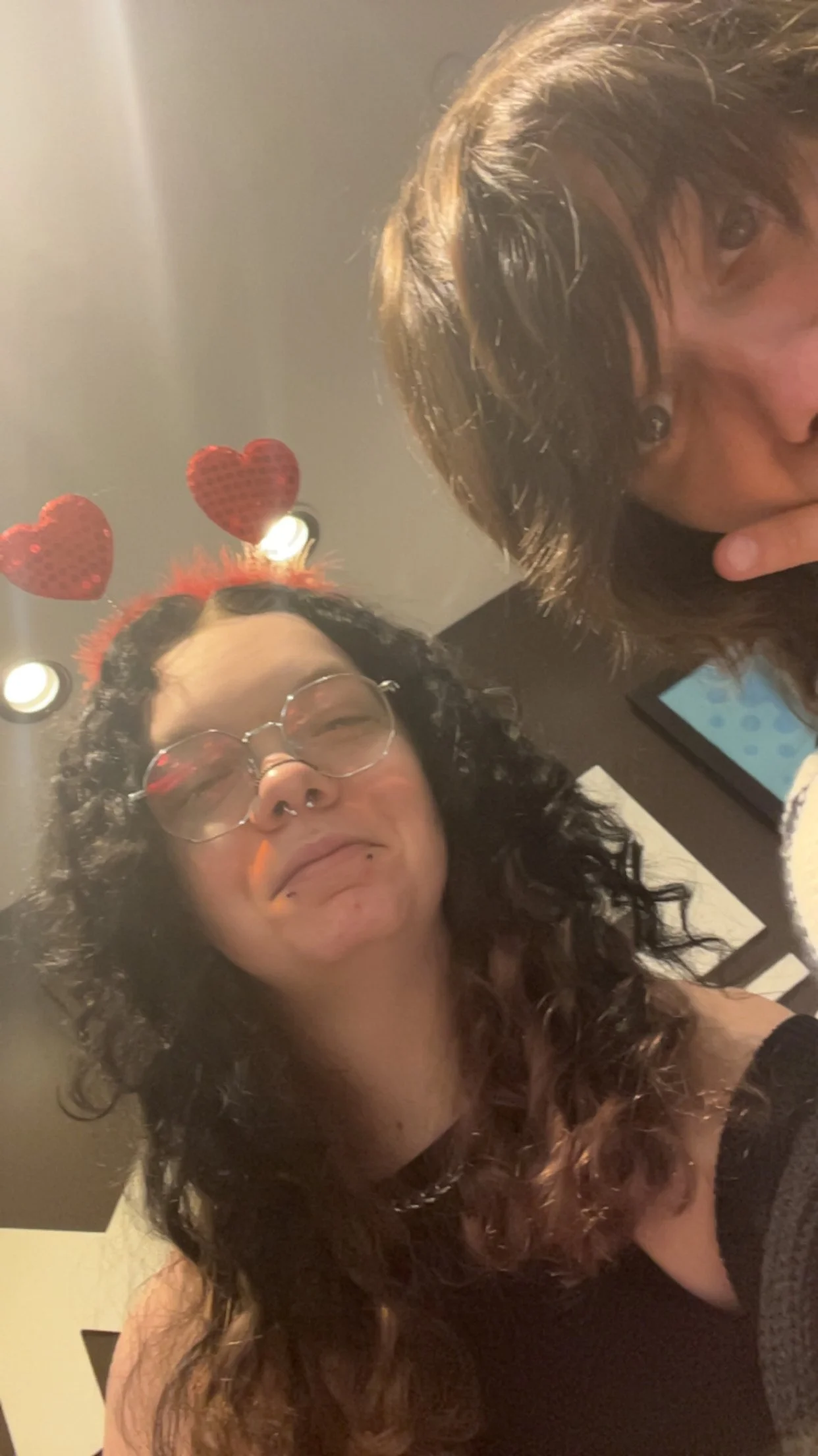 Two women, one with curly dark hair and wearing glasses, are taking a selfie indoors. One woman has a bow with red hearts on her head. The woman who is taking the selfie has dyed red hair. The photo appears to be taken in a casual environment.