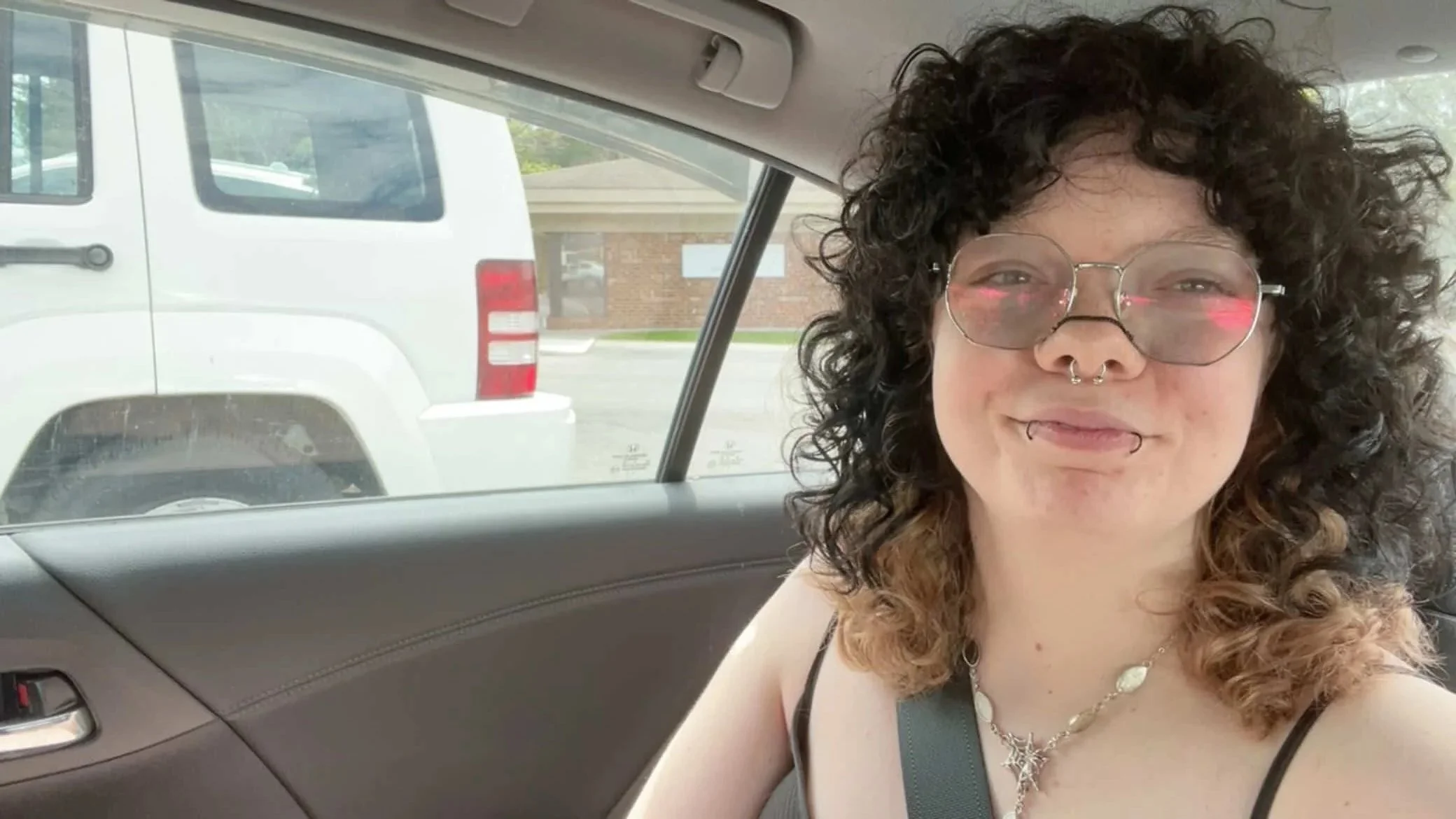 A woman with curly brown hair, wearing glasses and jewelry, smiling while sitting in a car.