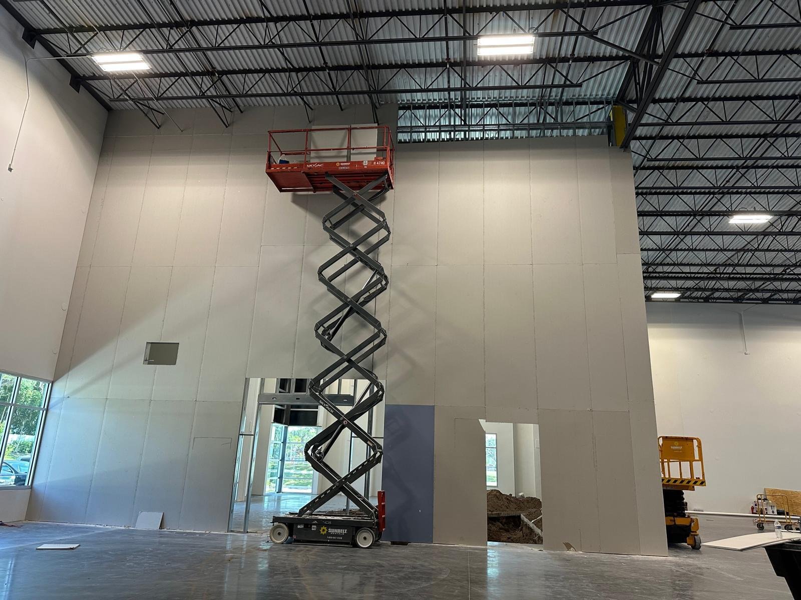 Construction inside a building with unfinished walls, a scissor lift reaching near the ceiling, and large windows letting in natural light.