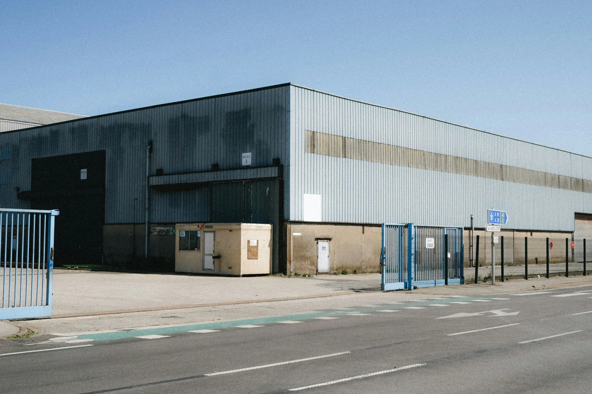 Industrial warehouse building with metal siding, a large sliding door, two smaller doors, and a sign indicating hall sections. Foreground features a parking lot with striped spaces and light blue fencing. Clear blue sky overhead.
