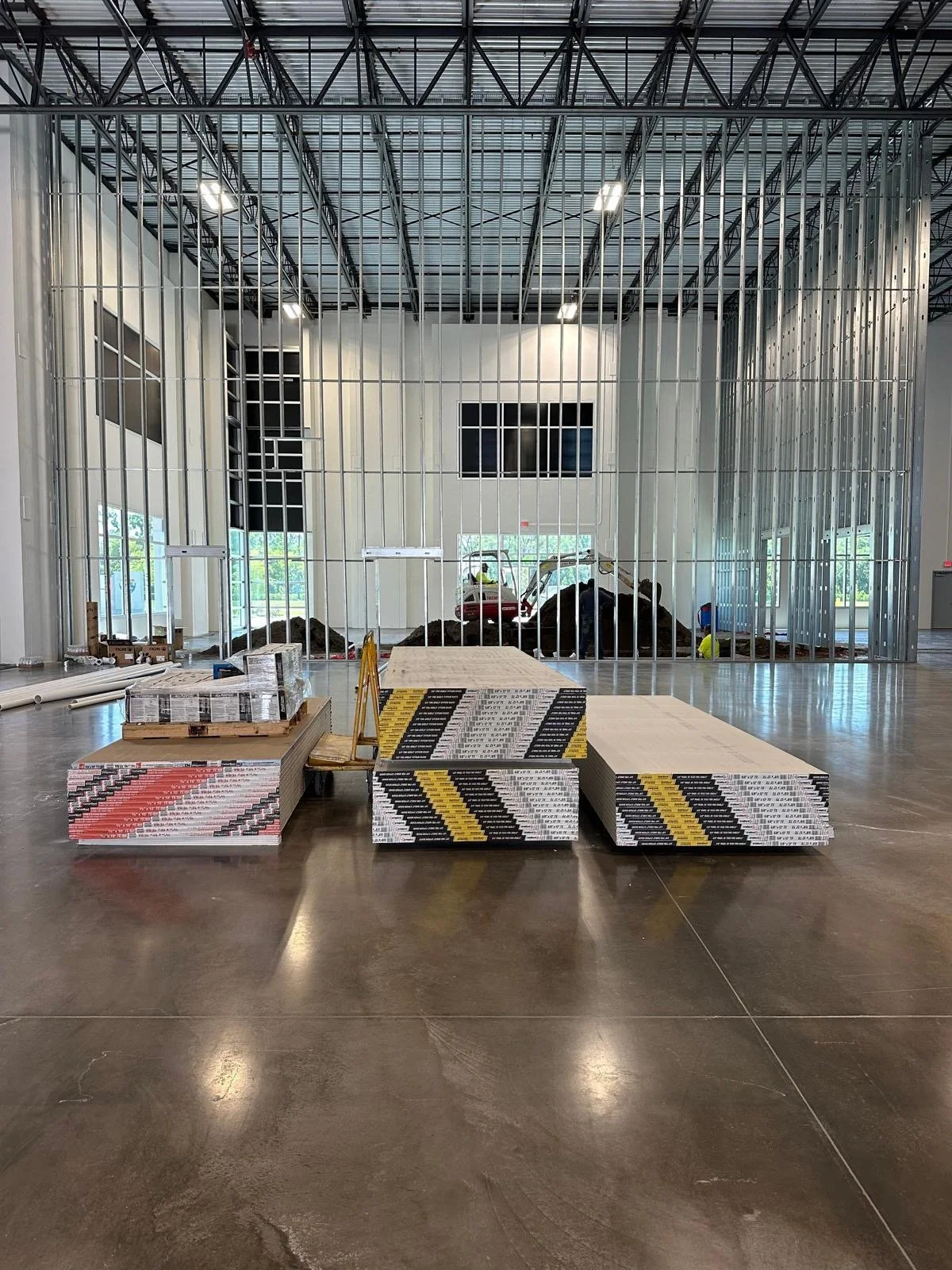 Construction site inside a building with metal framing for a wall or partition, construction materials in the foreground, and compact excavator at back, with debris and dirt on the ground.