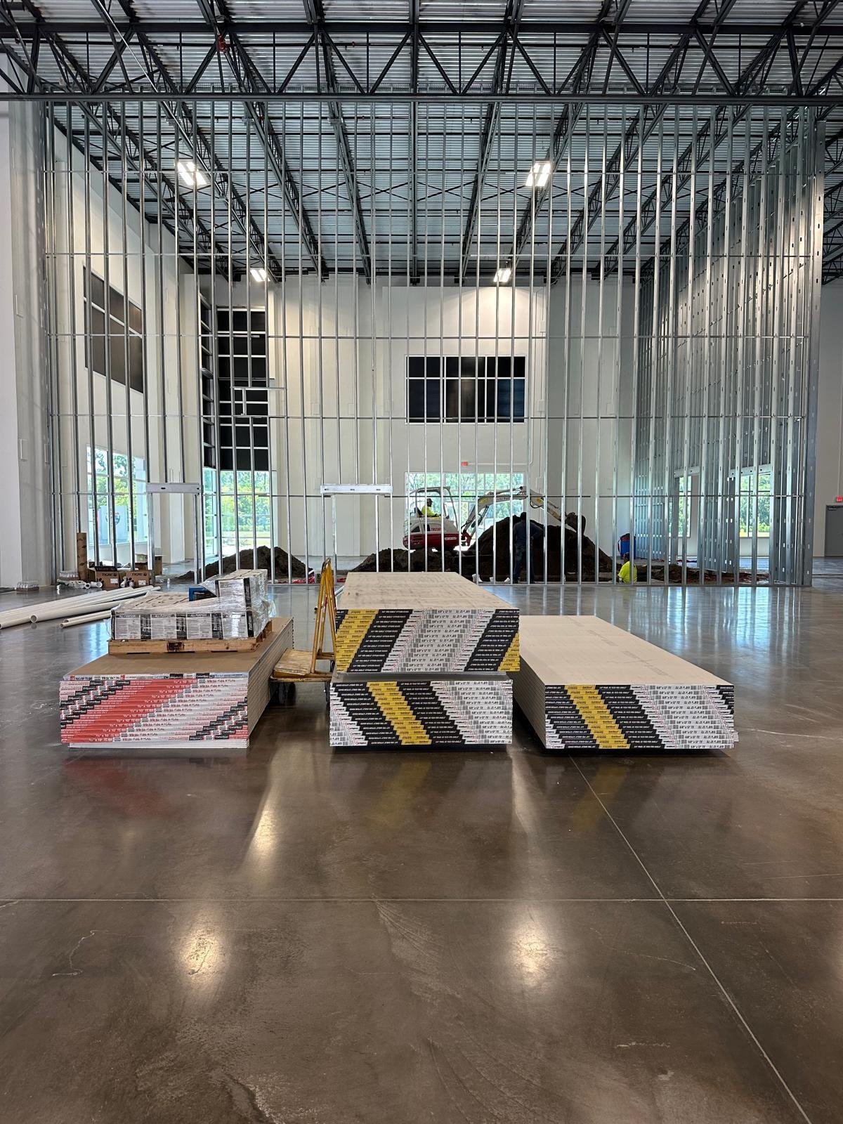 Construction materials and stacks of wall panels are arranged in an indoor space with a large, open area and high ceiling. Behind the materials, a metal framed wall is under construction, with construction tools, workers, and exposed terrain visible through the frame.