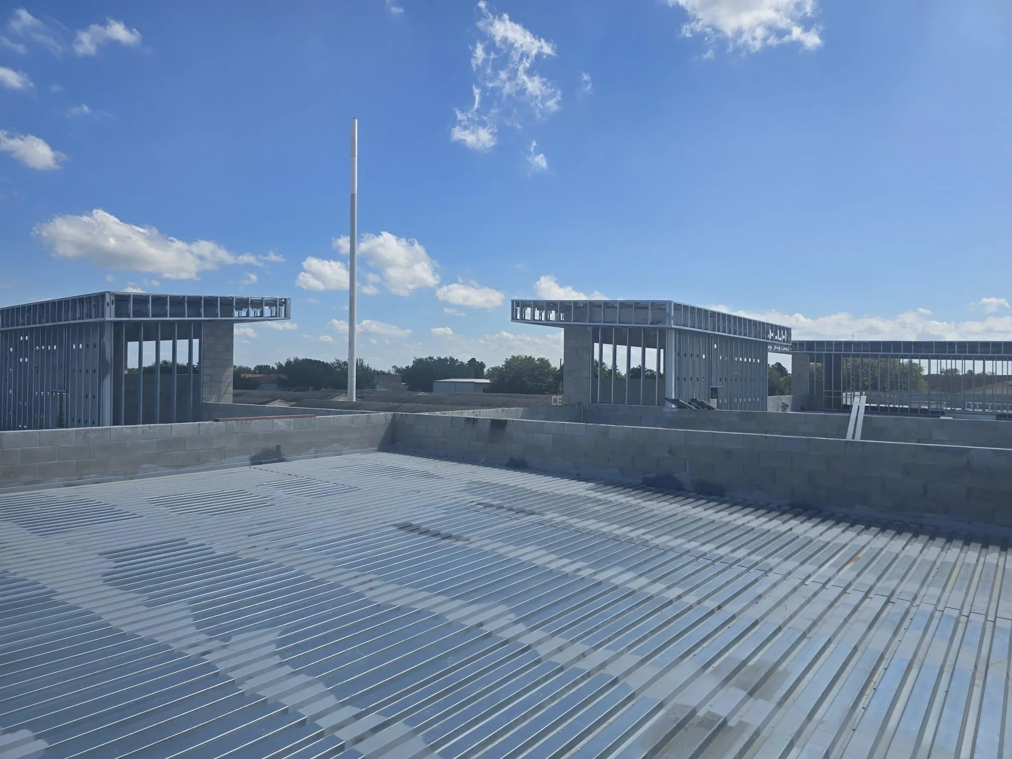 Rooftop with metallic solar panels, a low brick wall, and building structures with glass and metal, under a blue sky with some clouds.