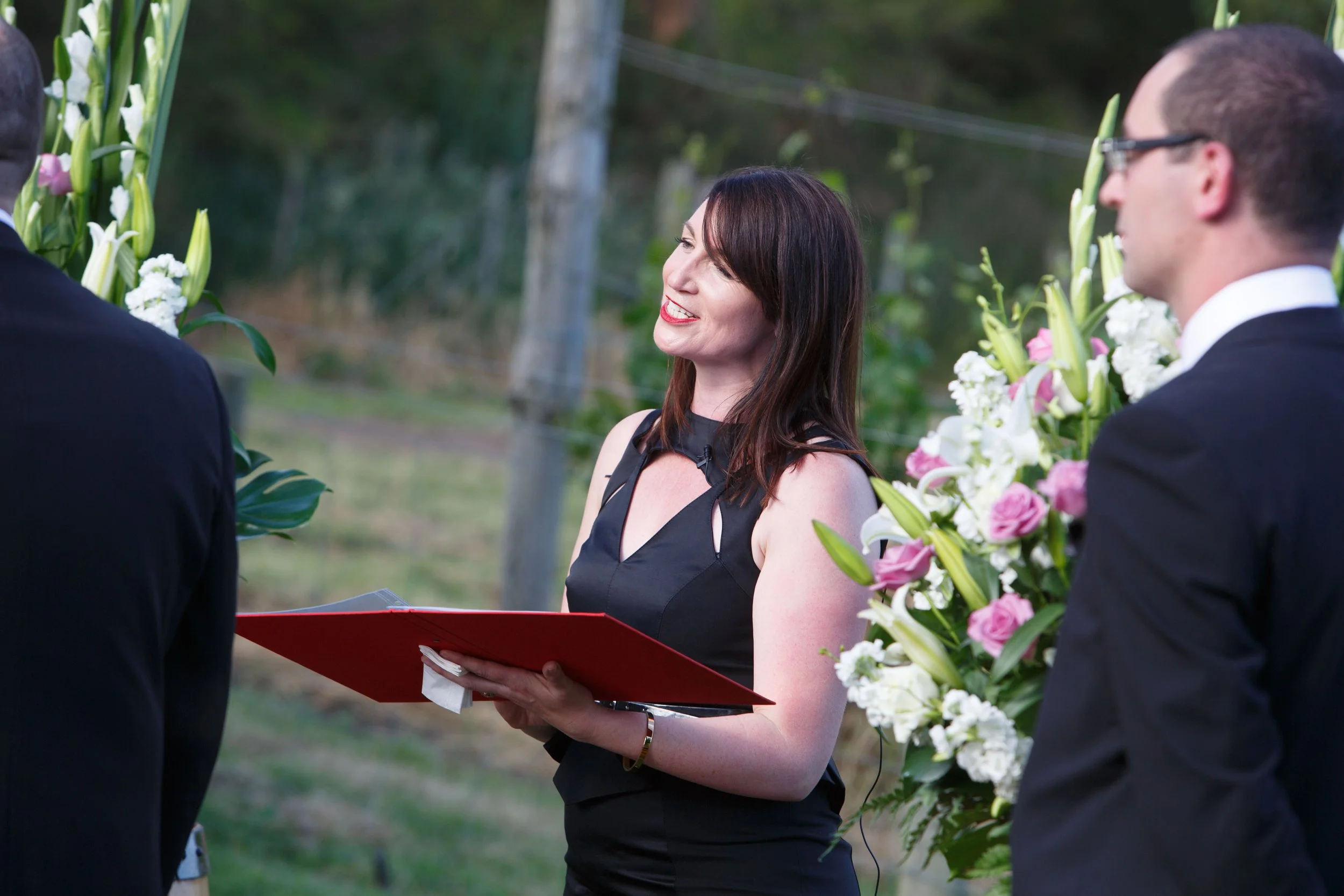 A woman in a black dress holding a red folder and smiling during an outdoor wedding ceremony, with men in suits and floral arrangements visible.