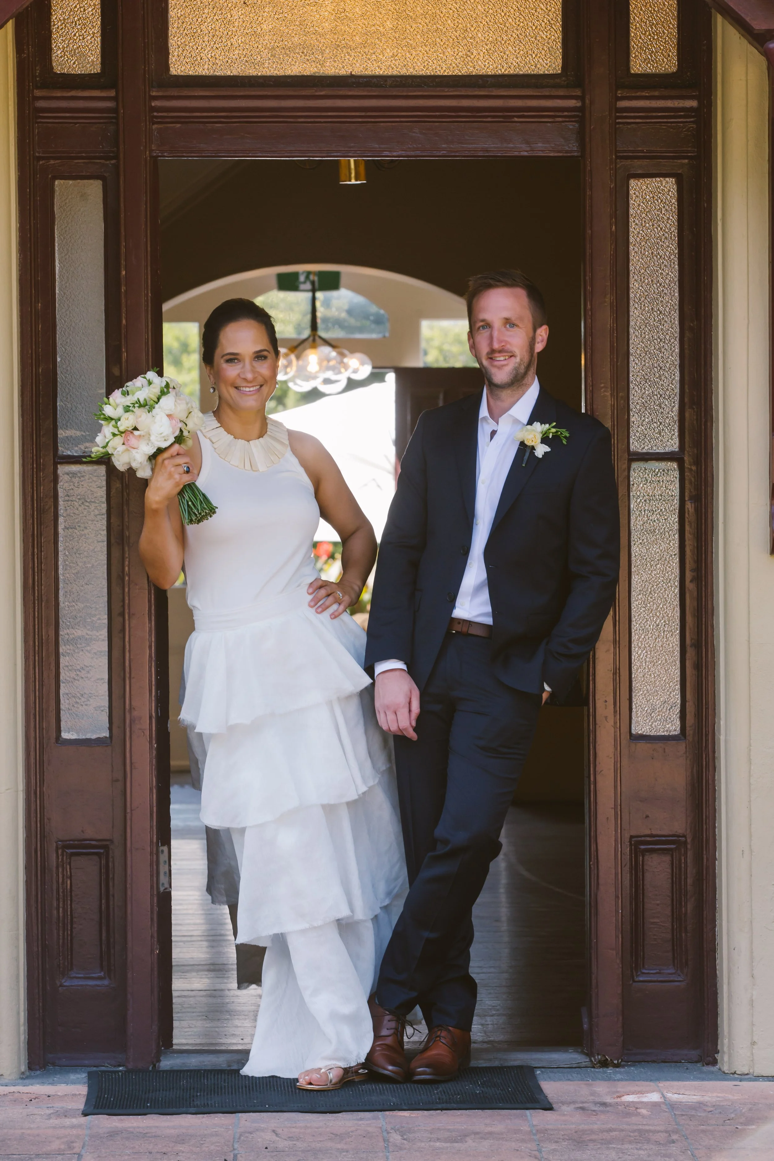 A newlywed couple stands in the doorway of a building, smiling at the camera. The bride holds a bouquet of white and pink flowers, wearing a white dress with ruffles and a statement necklace. The groom wears a dark suit with a white shirt and boutonniere. The setting appears to be bright and cheerful, with natural light and greenery visible in the background.