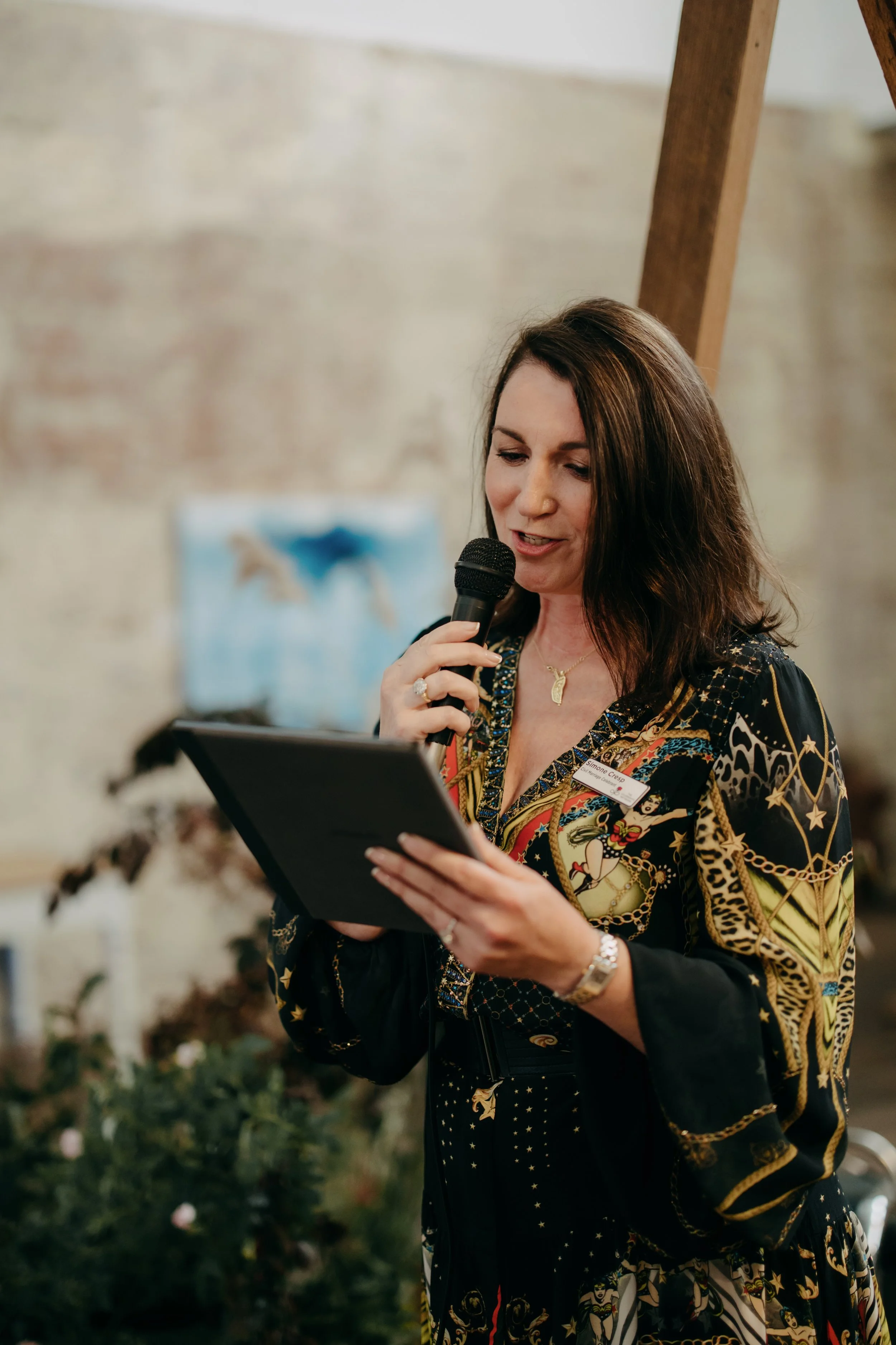 A woman with long brown hair, wearing a colorful, patterned blouse, holds a microphone and a tablet, standing in front of a decorated background with a Christmas tree and a painting of horses.