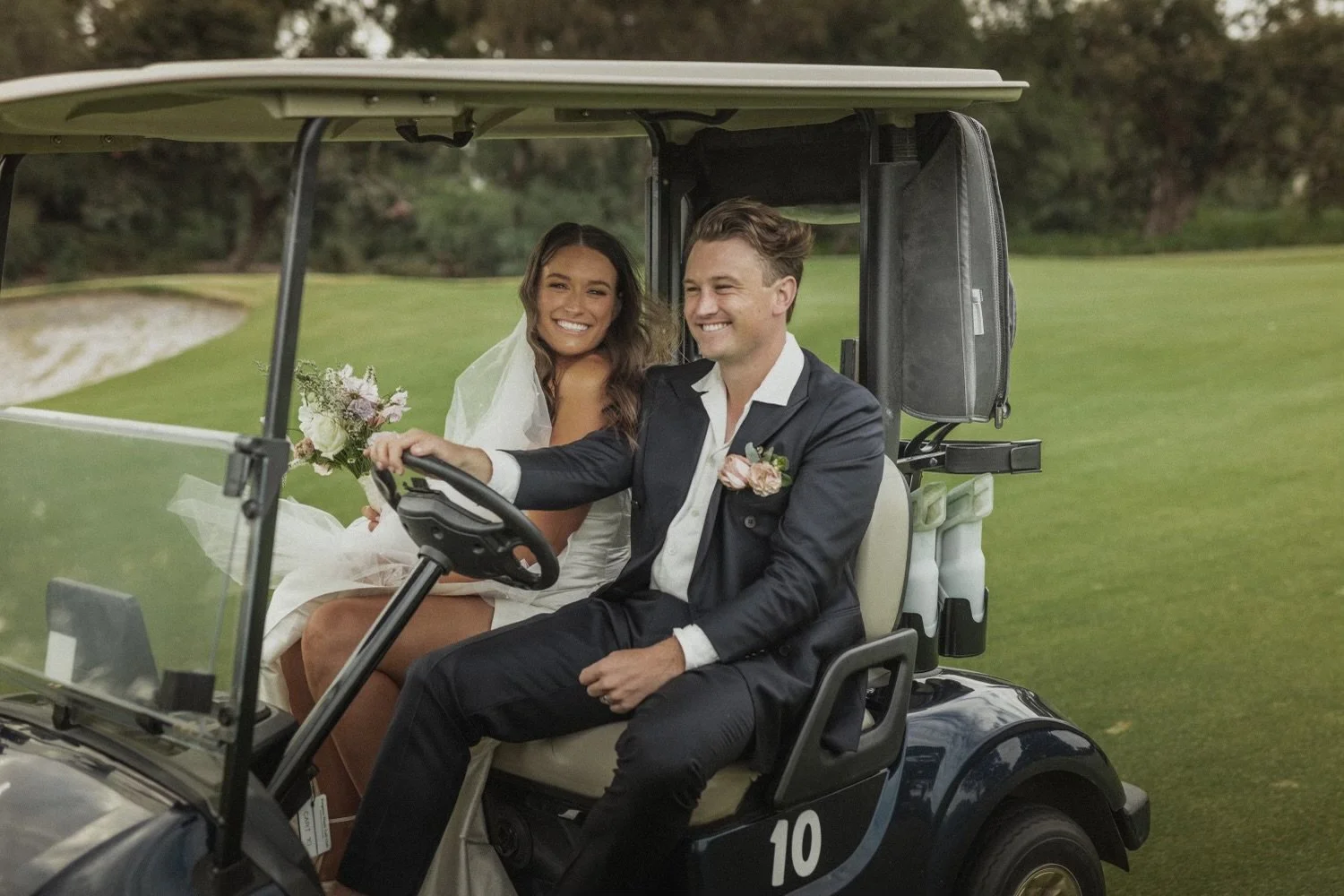 A newlywed couple sitting in a golf cart on a golf course, smiling and enjoying their day.