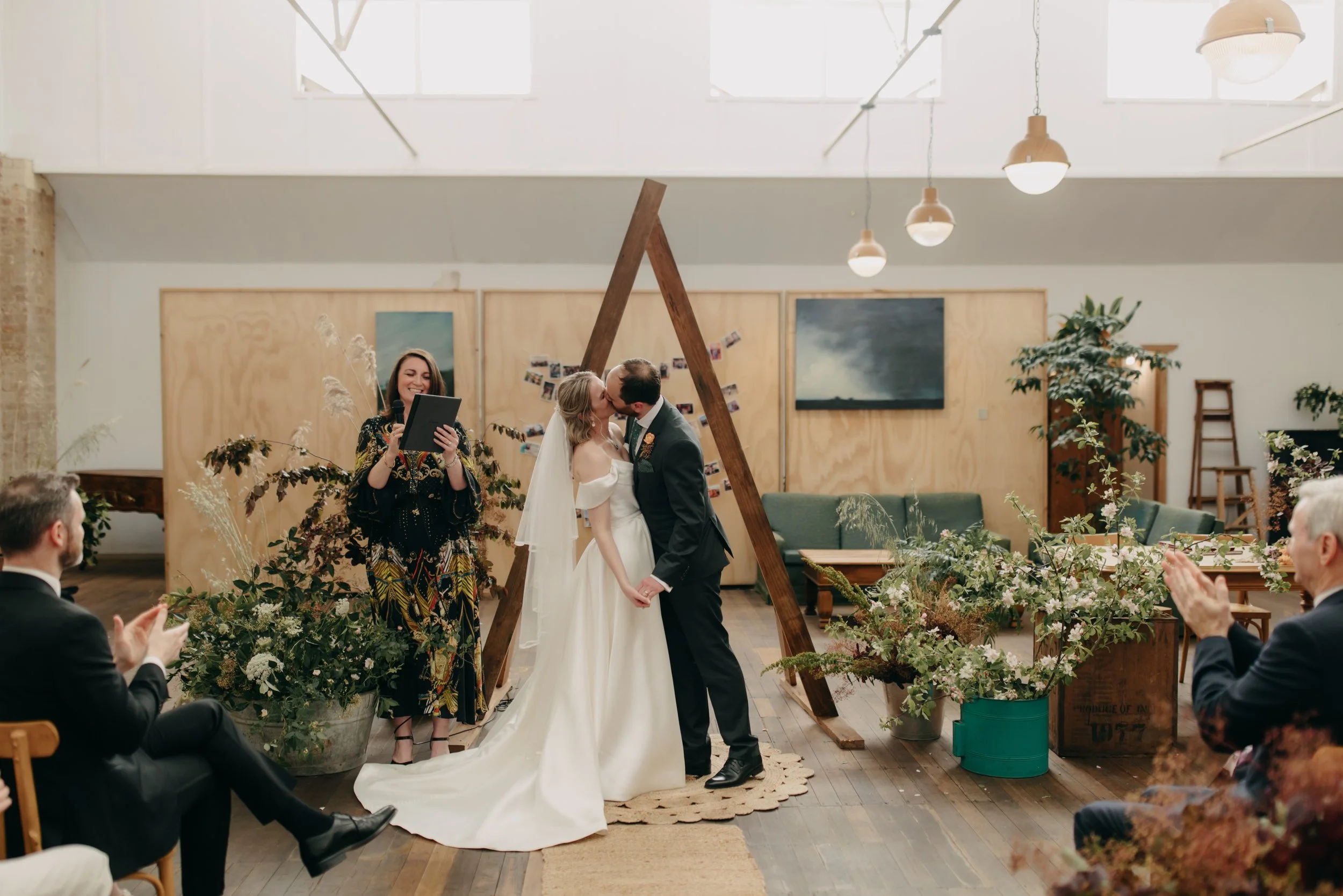 A couple shares a kiss during their wedding ceremony in a modern indoor venue decorated with green plants and flowers. Guests are seated and clapping, a woman officiant holds a microphone, and the couple stands on a small circular rug, with a wooden A-frame structure behind them.