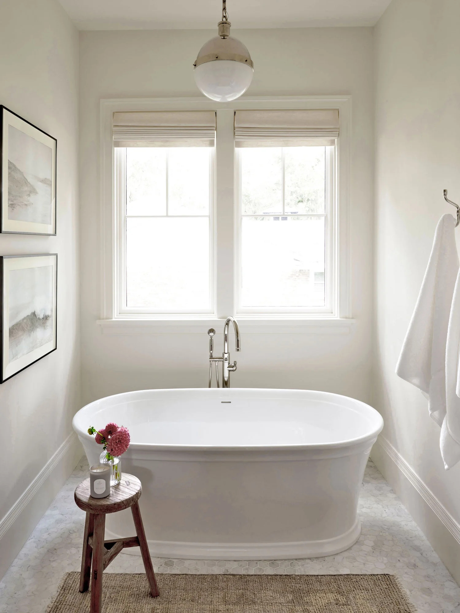 Bright bathroom with white bathtub, large window, and minimalist decor. A small wooden stool holds candles and flowers, with a framed artwork on the left wall and a towel hanging on the right.
