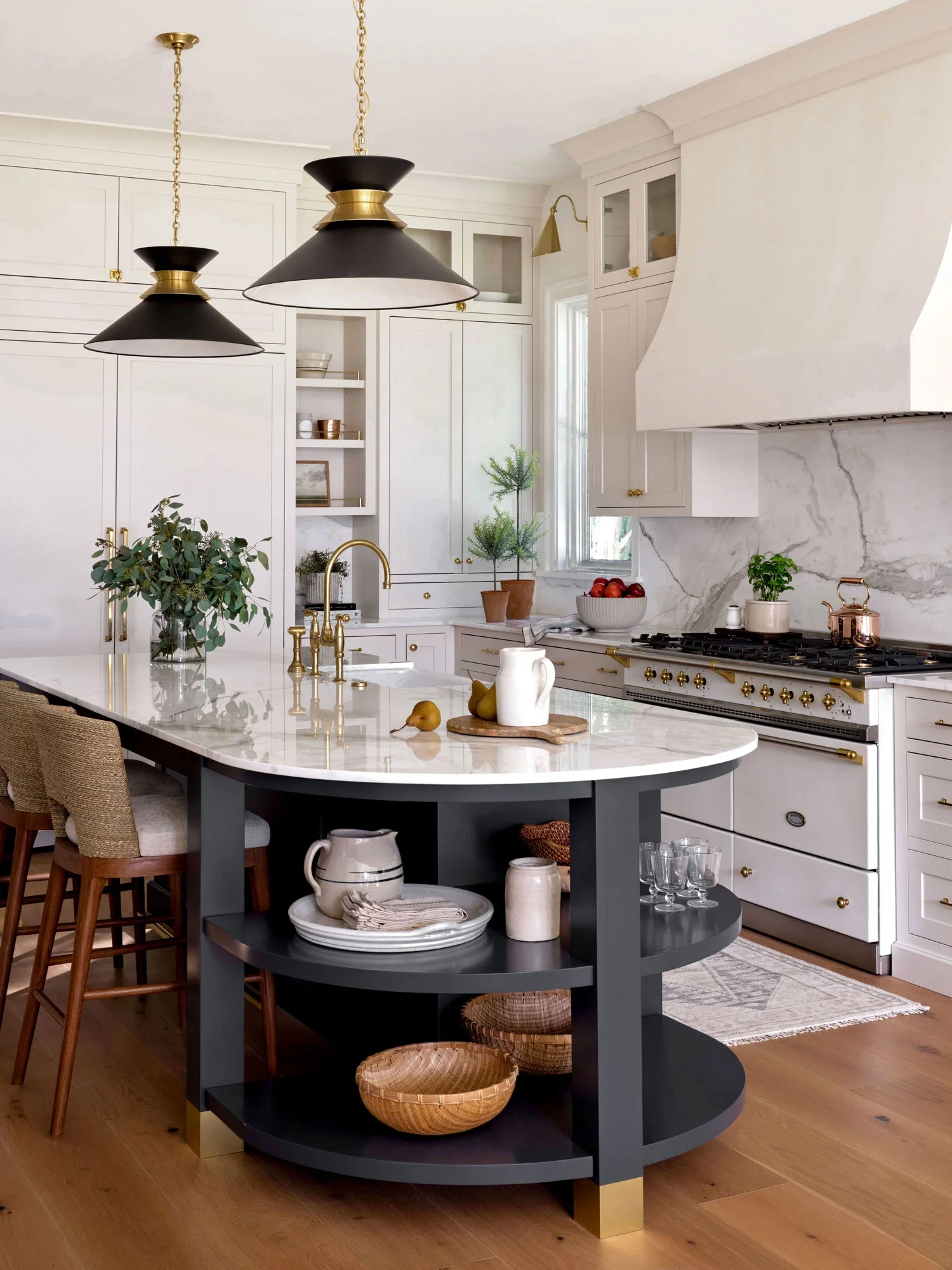 A modern kitchen with white cabinets, a marble backsplash, and black and gold pendant lights above a dark gray kitchen island. The island has a marble countertop, gold fixtures, and open shelves with pottery and woven baskets. The kitchen features gr