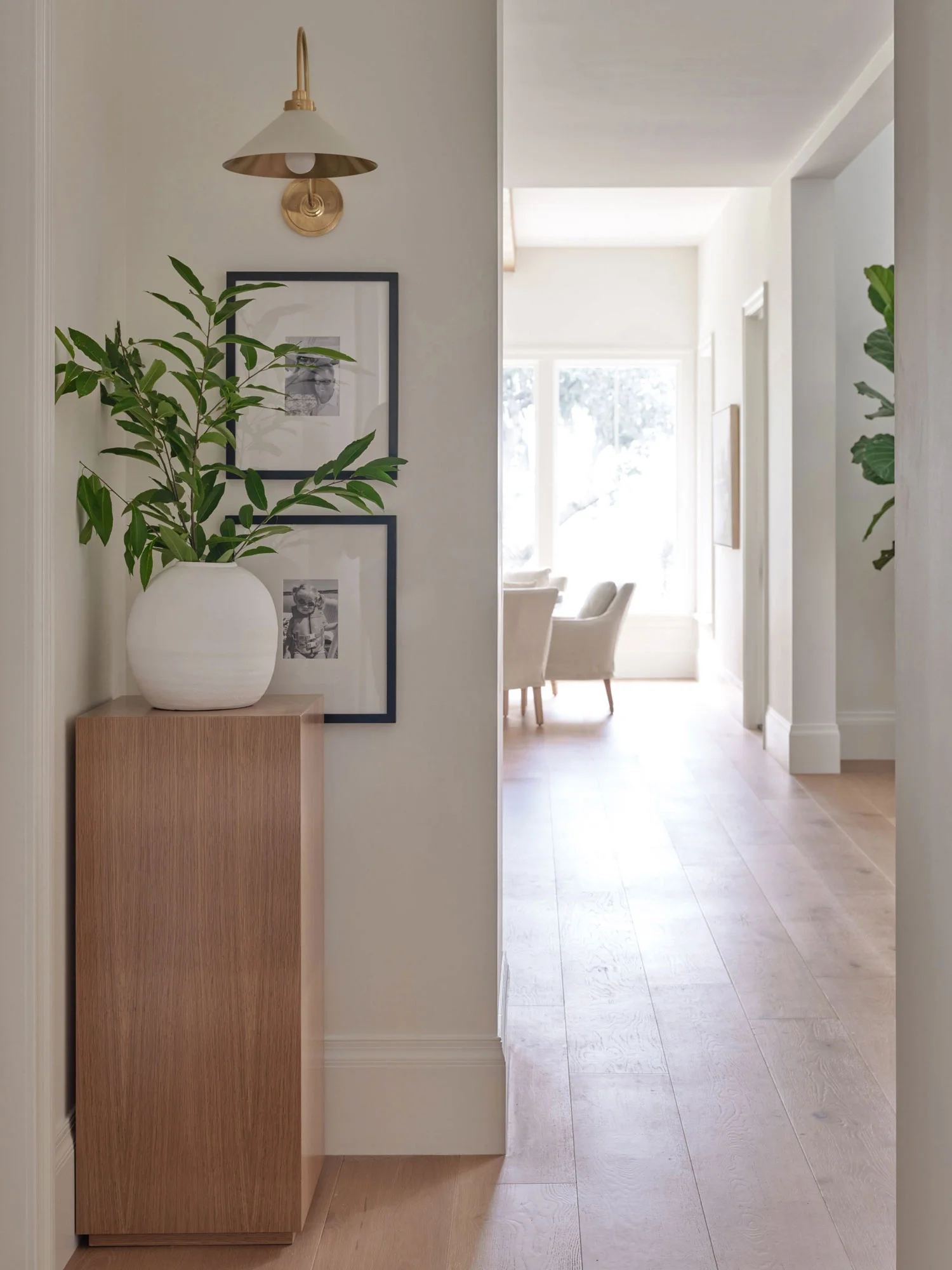 Interior view of a hallway with a potted plant on a wooden stand, framed photographs, a wall-mounted lamp, and a bright room with armchairs visible in the background.