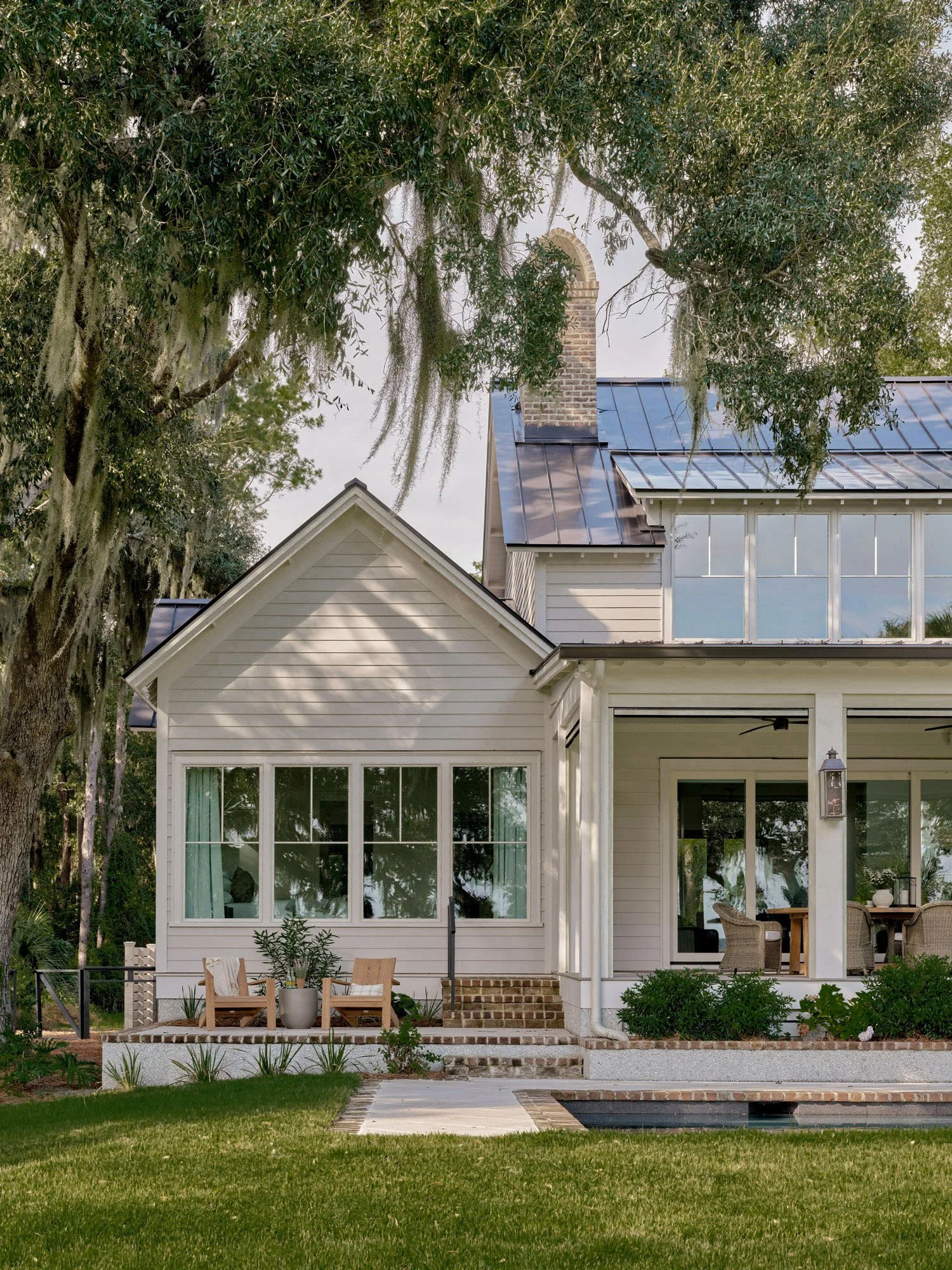 A two-story house with a white exterior and large windows, featuring a screened porch and a sloped roof with brick and metal elements. The house is surrounded by a well-maintained lawn, plants, and tall trees with hanging moss.