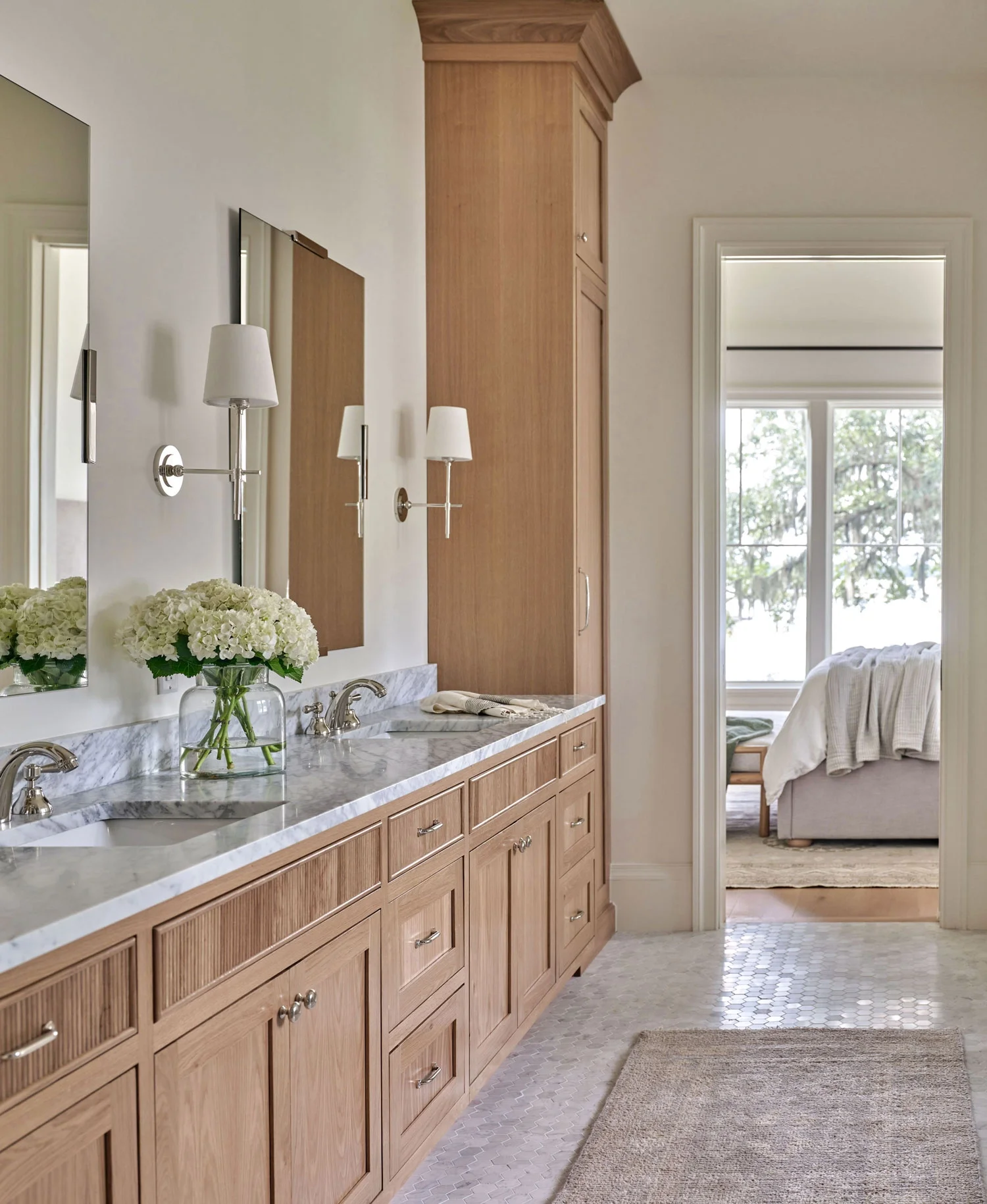 A bathroom with a long wooden vanity, marble countertop, and two sinks. Above the vanity, there are two wall-mounted lamps and a mirror. A vase with white flowers and a soap dispenser are on the countertop. There is a wooden cabinet with drawers and 