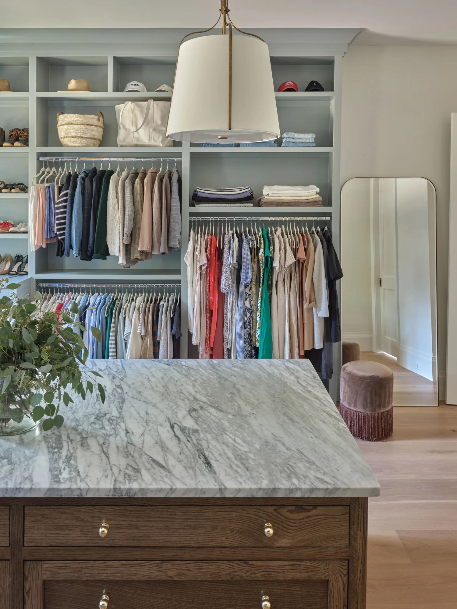A walk-in closet with light and dark clothing, hats, and bags on built-in shelves. A large white lamp hangs from the ceiling. A marble-topped dresser is in the foreground, and a small brown ottoman is near a mirror.