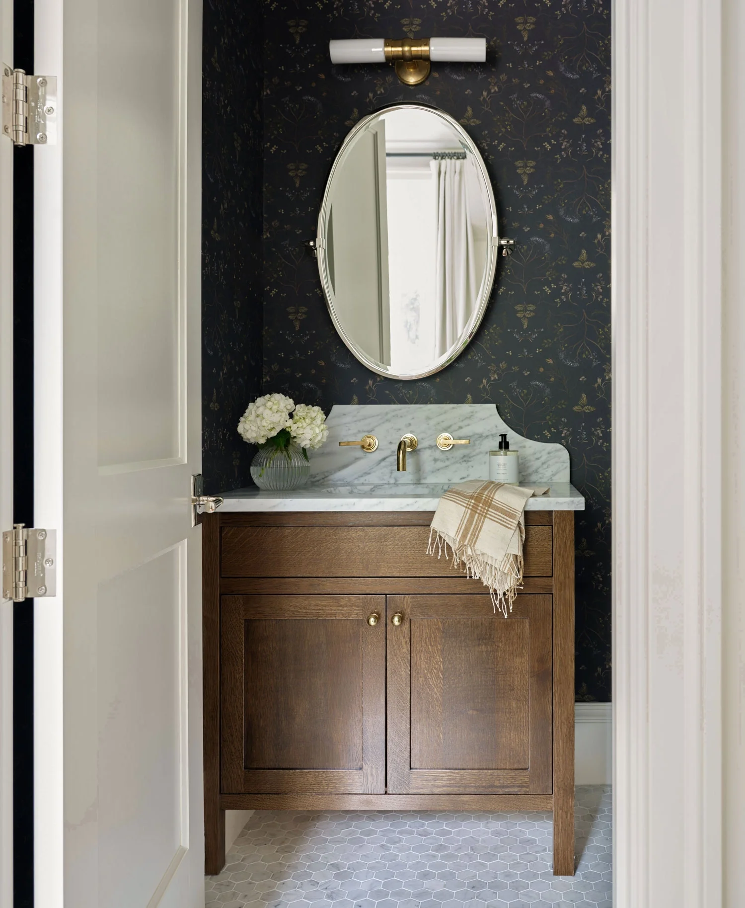 A bathroom vanity with a marble countertop, a round mirror, a gold faucet, a flower vase with white flowers, and a hand towel. The vanity is made of wood with gold knobs, and there is a wall-mounted light fixture above the mirror. The wall has dark f