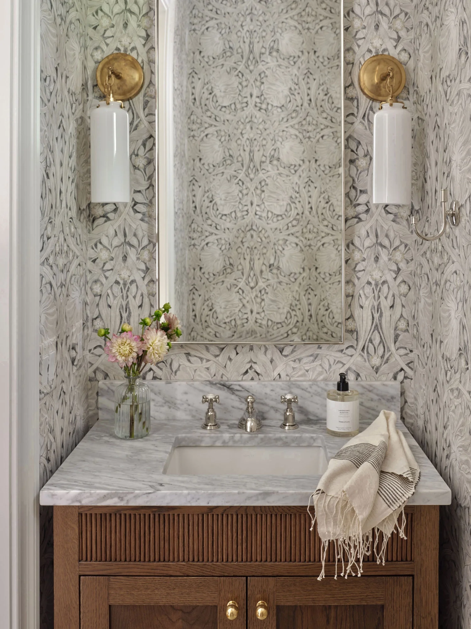 A bathroom vanity with a marble countertop, a rectangular sink, and a wooden cabinet with gold knobs. There is a glass vase with pink and white flowers, a soap dispenser, a rolled towel, and a hand towel on the counter. The wall above has patterned w